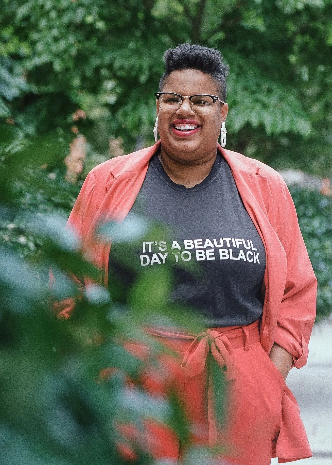 Katherine—a Black woman with a mohawk—is wearing an orange suit and a t-shirt that says “It’s a Beautiful Day to be Black” while standing behind a green bush