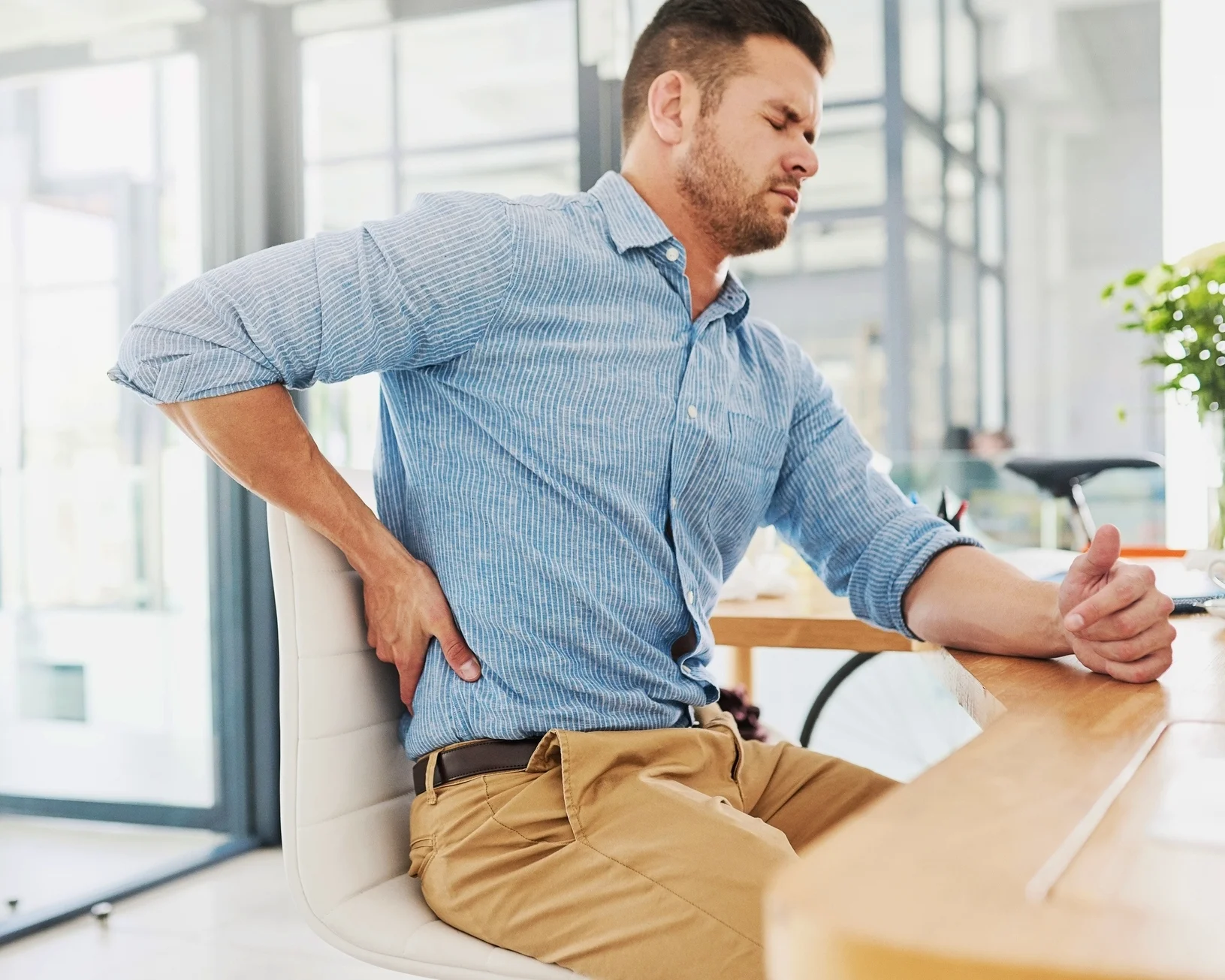 Office worker sitting at his desk with his eyes closed, holding his lower back in pain.