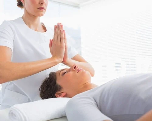 Woman relaxing on a treatment table while a Reiki practitioner gently holds her hands above the woman's face during an energy healing session.