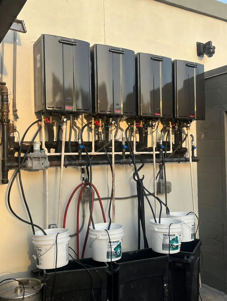 Multiple black electronic control units mounted on a wall, with tubes and wires connected to buckets and black containers below, possibly for a hydronic or glycol system.