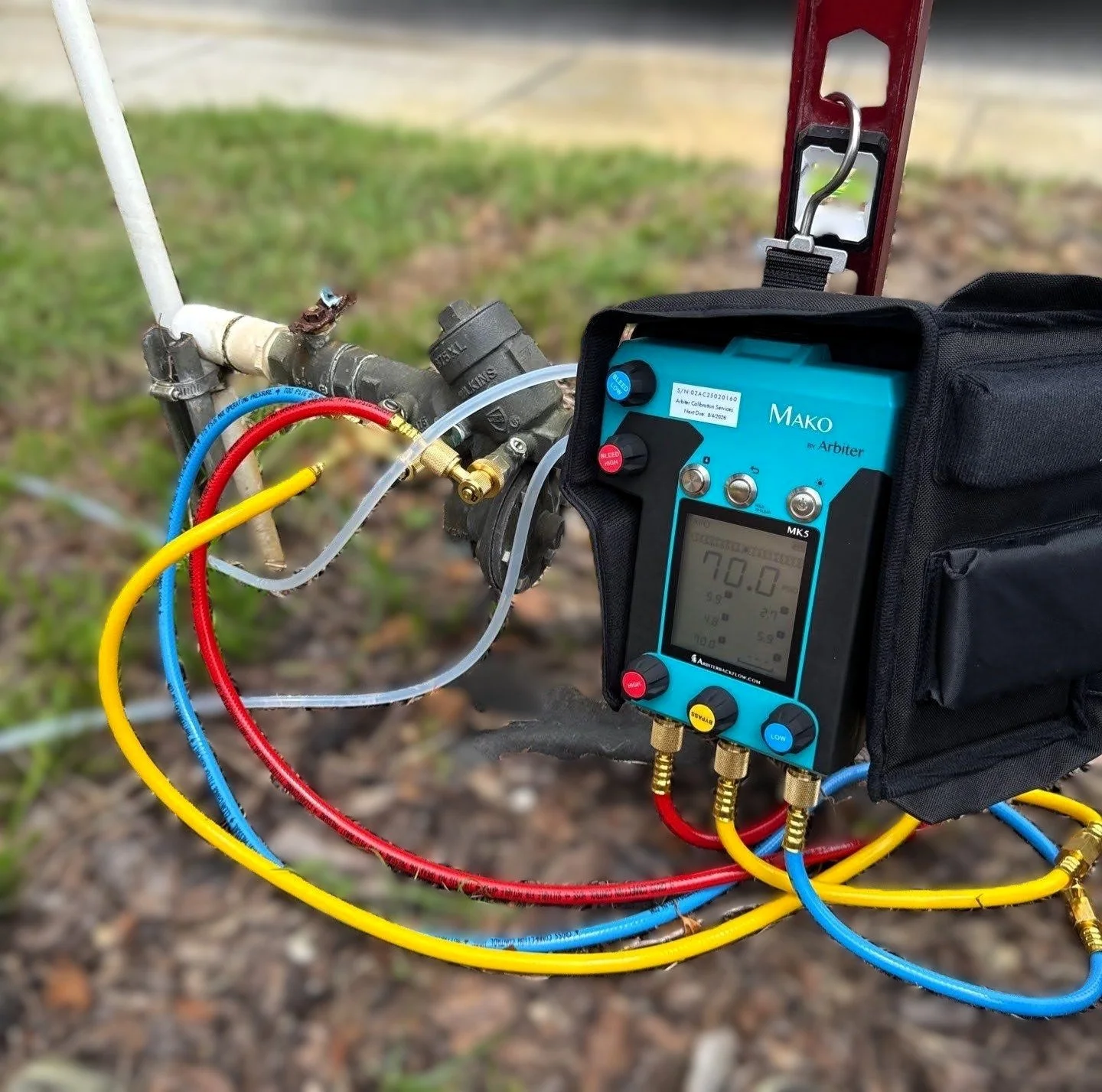 A digital manifold gauge used for HVAC or refrigeration system testing, connected to pipes with colored hoses and displaying readings on a digital screen.