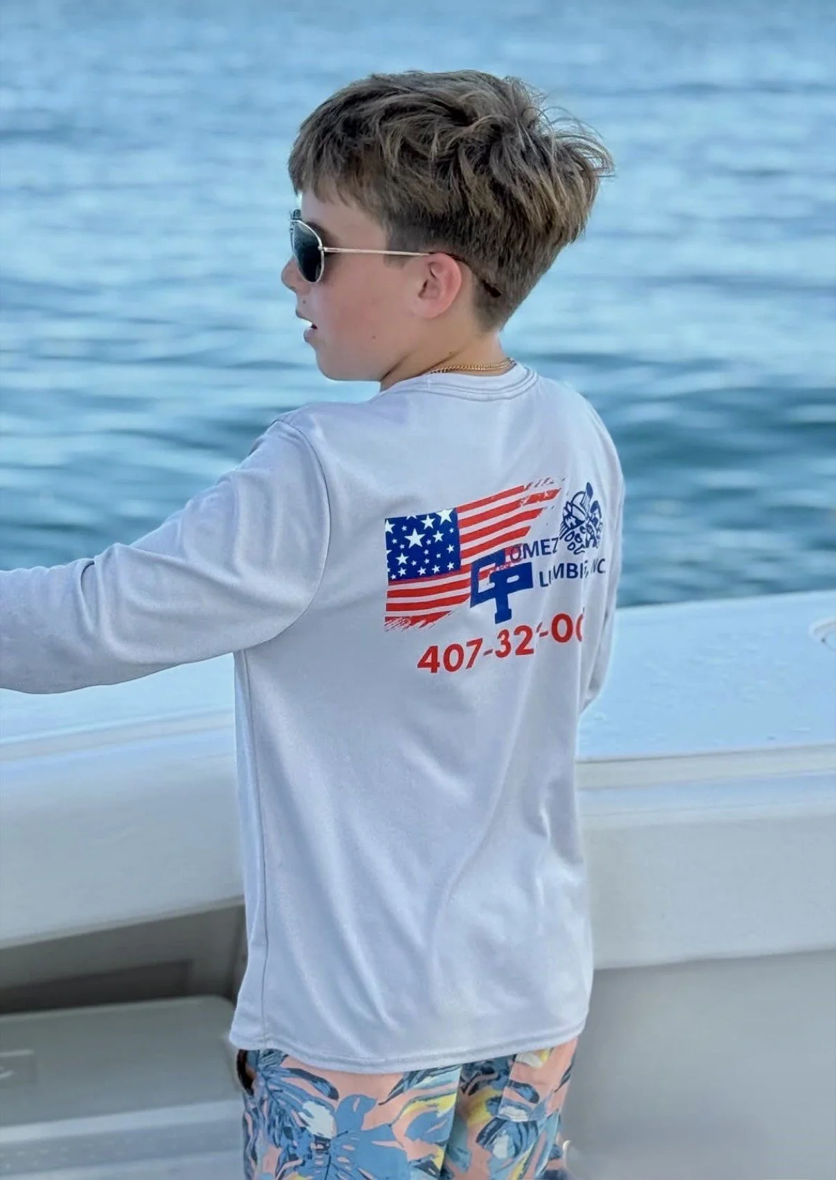 A young boy with brown hair wearing sunglasses, a white long-sleeve shirt with American flag and logo, and colorful shorts, standing on a boat near water.