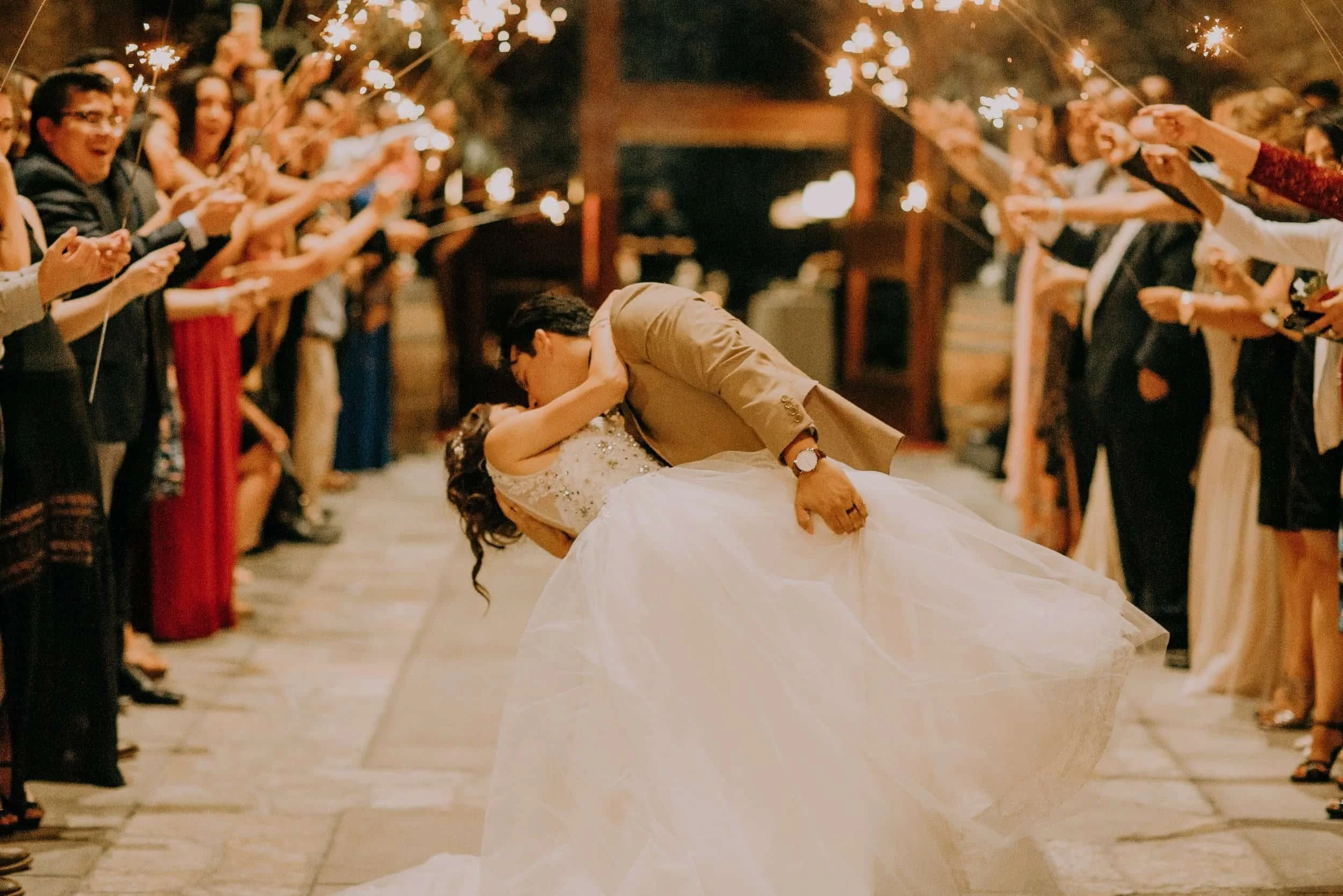A bride and groom sharing a kiss while dancing at their wedding reception, surrounded by people holding sparklers in a festive outdoor setting. They learned by taking wedding dance lessons online.