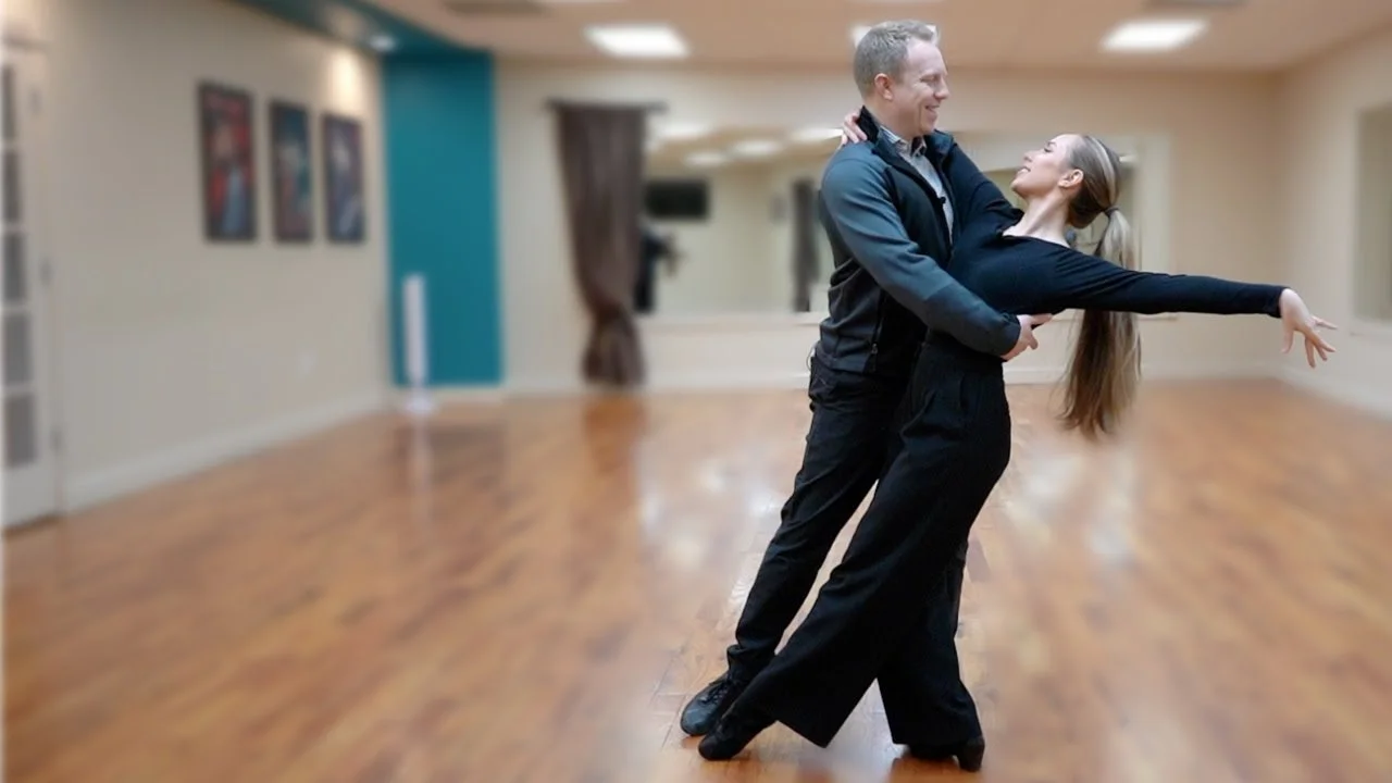 A man and woman practice wedding dances, perfecting their first dance in an empty studio with wooden floors and mirrors.