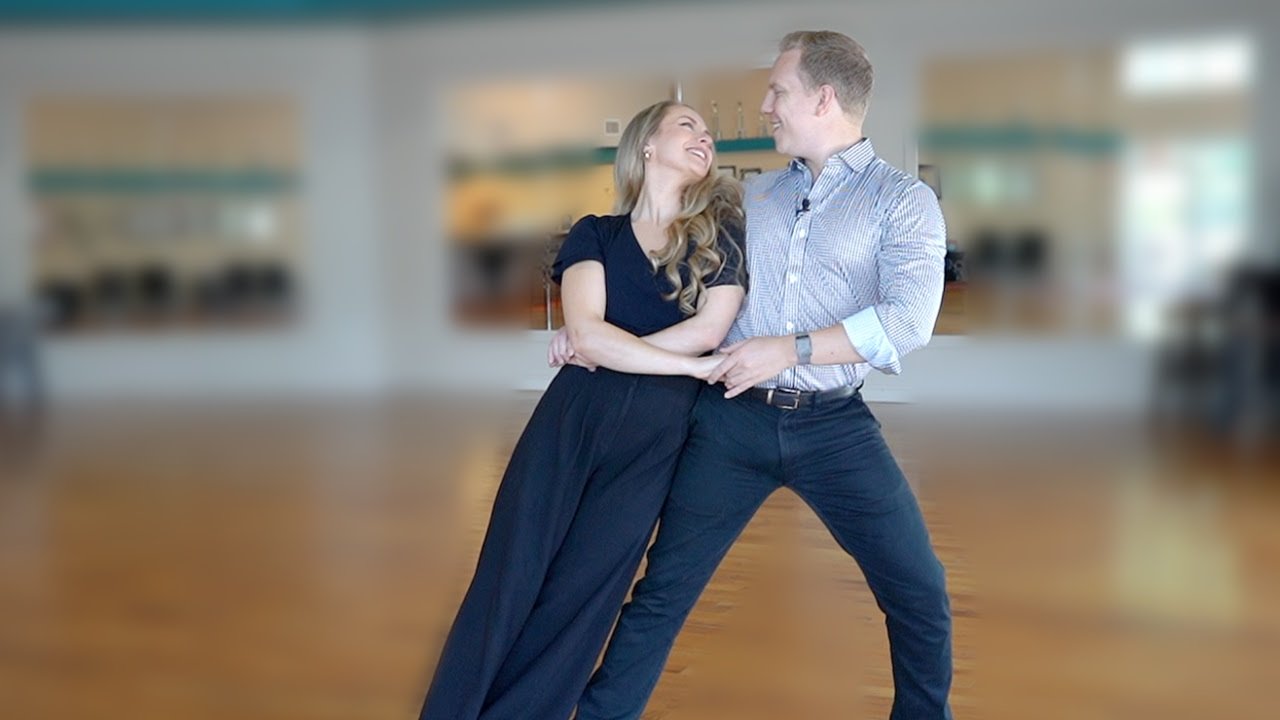 A smiling couple practices their wedding dances in a bright, spacious studio with wood floors and large windows.