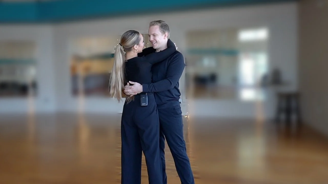 Two people in black outfits practice a wedding dance hold in a spacious, bright room with wooden floors and large mirrors.