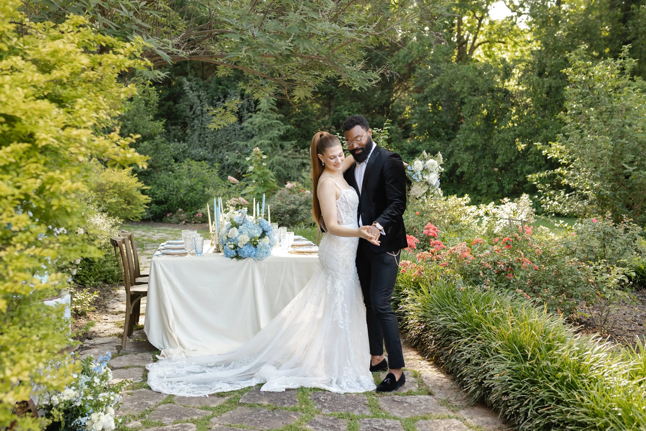 A bride and groom dancing outdoors at a wedding reception with a decorated table and lush greenery in the background.