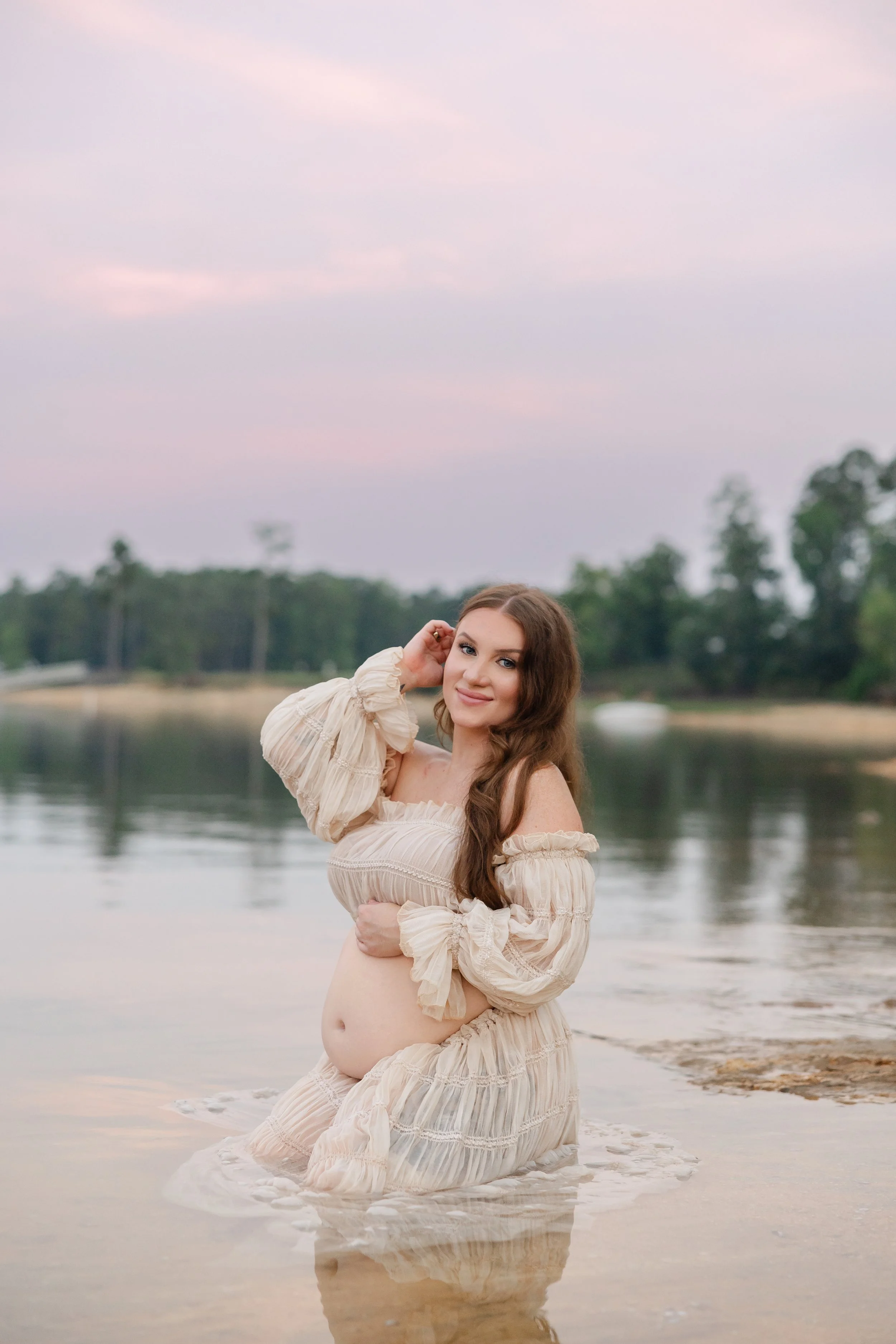 Pregnant woman in a flowing cream maternity dress kneeling in a lake at sunset during a lakeside maternity session in Appling, Georgia.