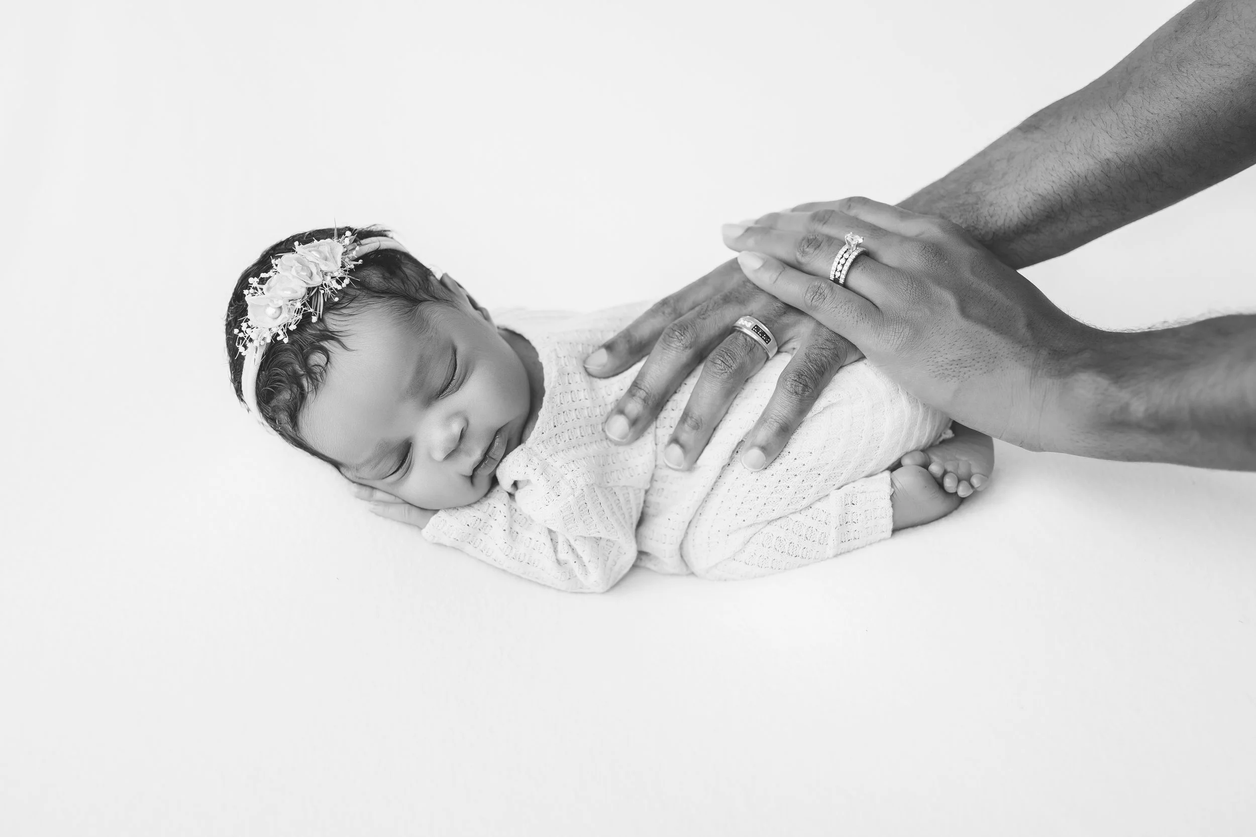 A newborn baby sleeping on a soft surface, with an adult’s hands gently touching the baby’s back. The baby is wearing a floral headband and a long sleeve outfit, and the adult’s hands are adorned with rings.