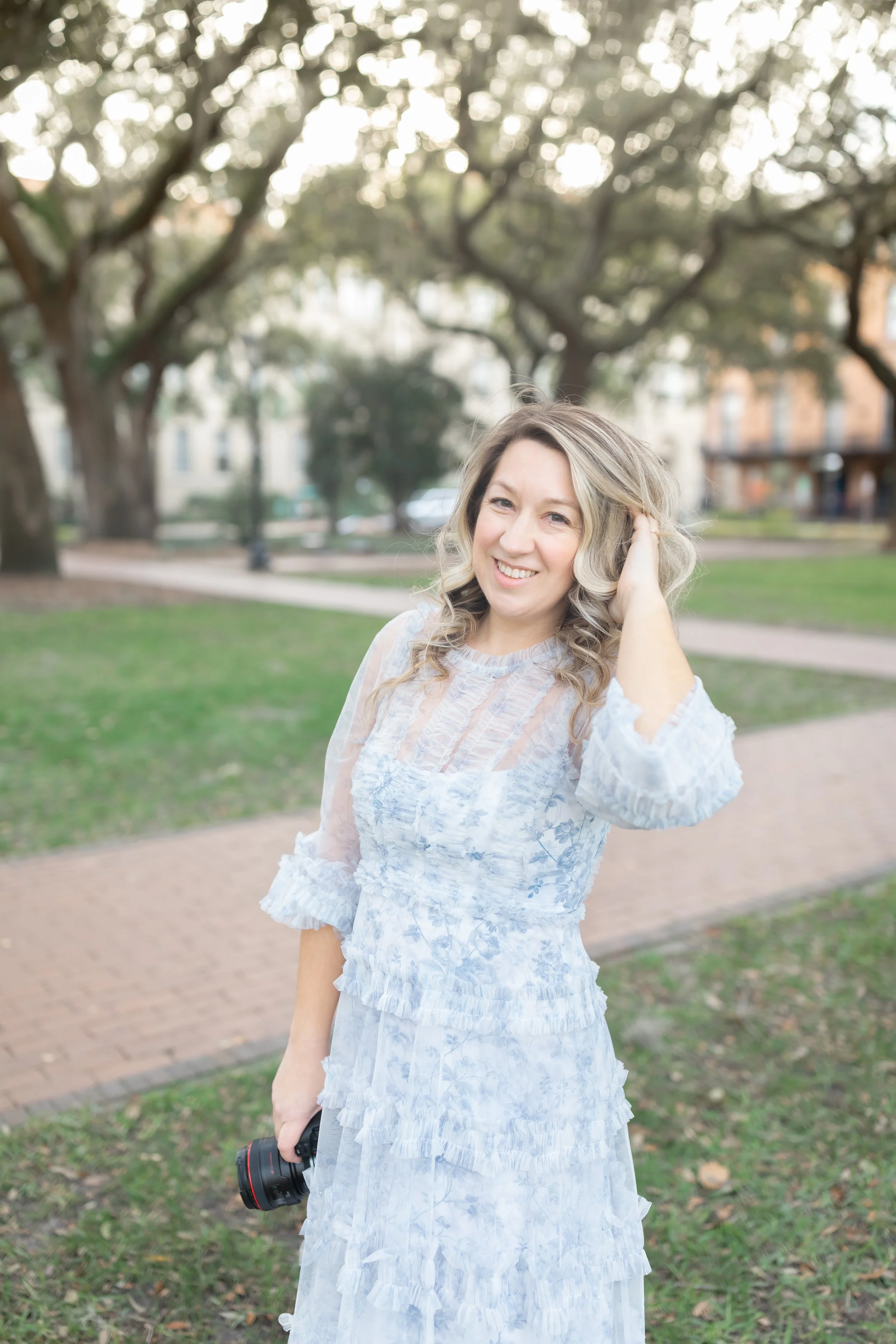 A smiling woman with blonde curly hair wearing a white lace dress, holding a camera in her right hand, standing outdoors in a park with trees and a brick walkway in the background.