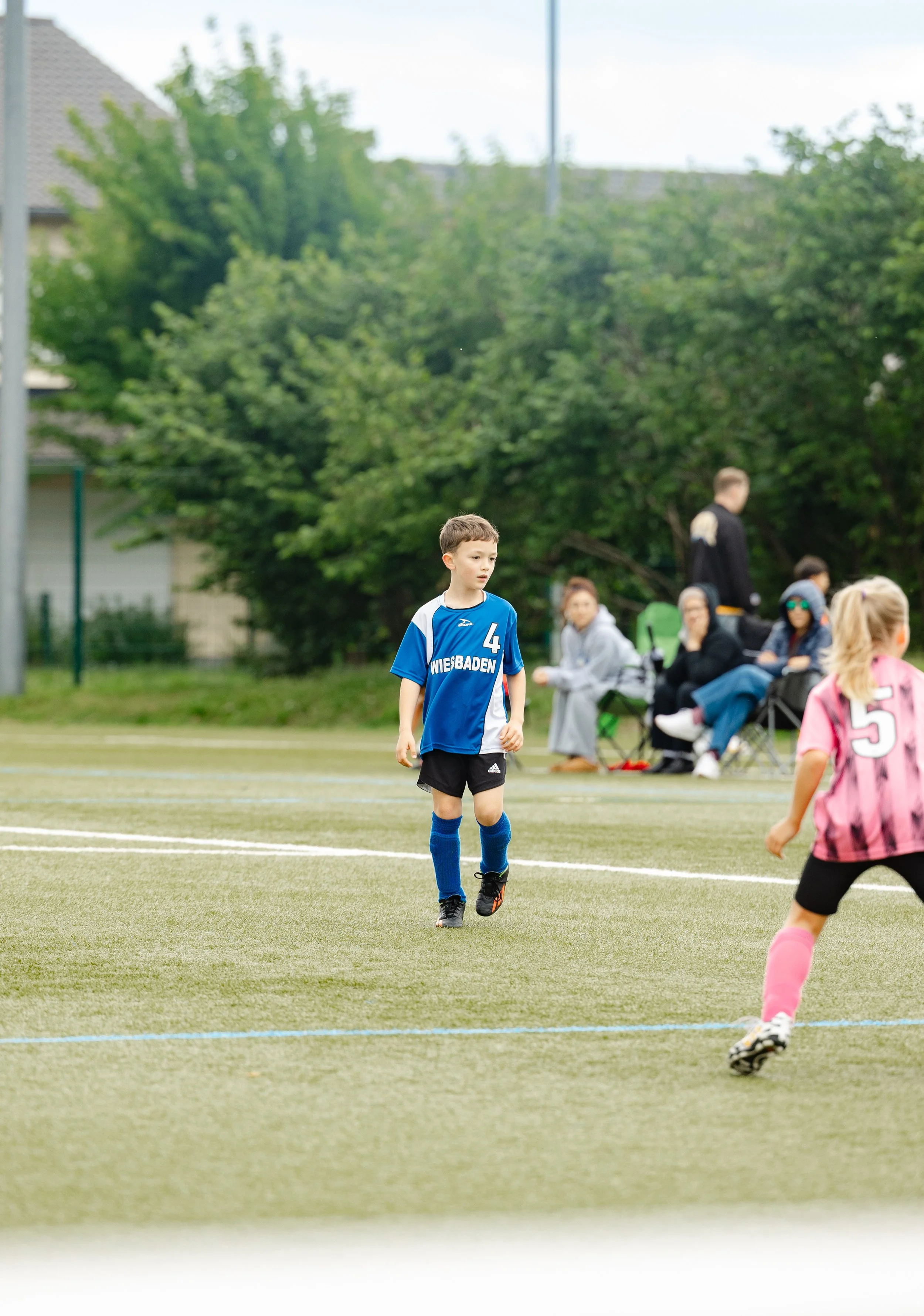 Children playing soccer on a field while spectators watch from benches in the background, with trees and residential buildings around.