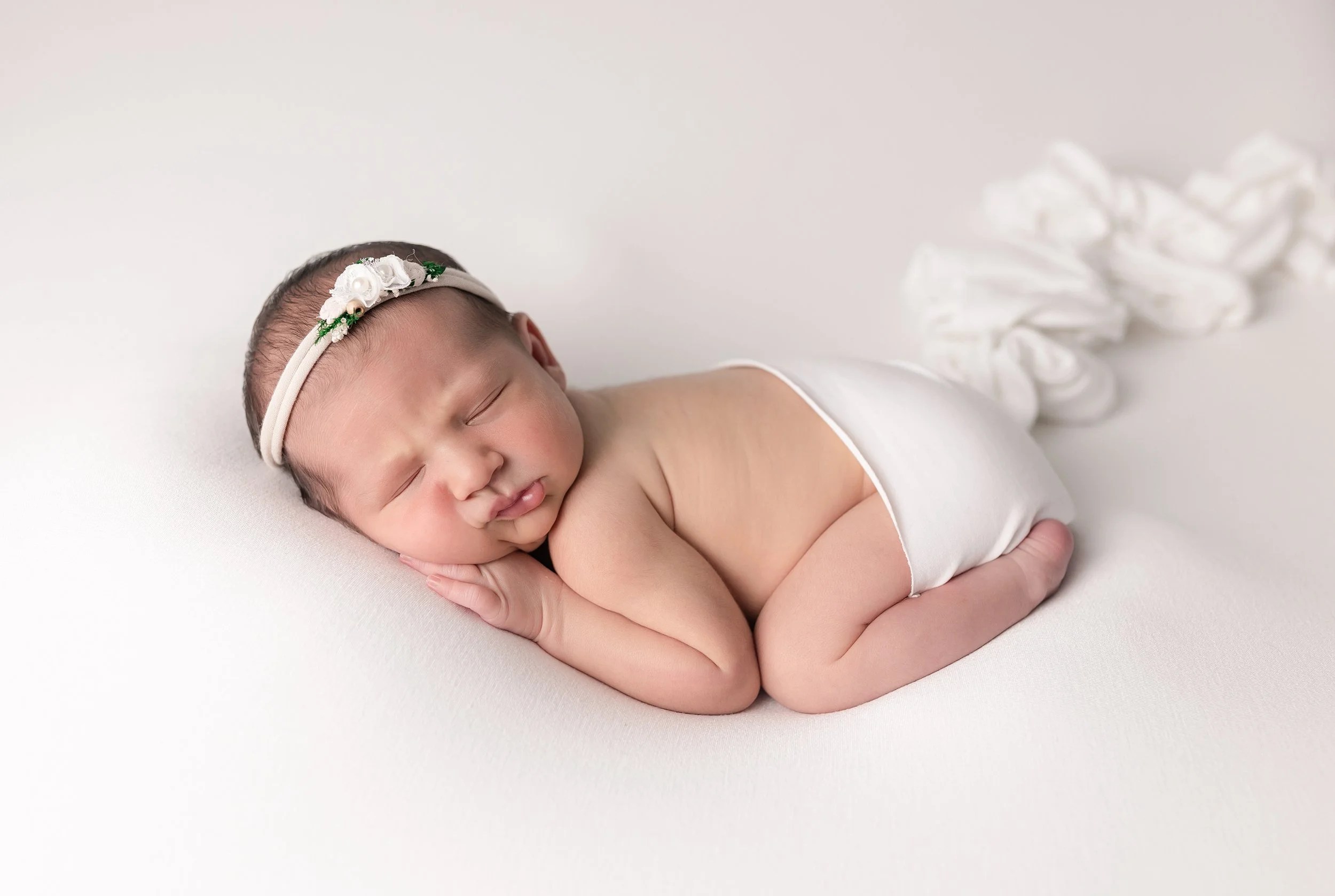 A newborn baby girl sleeping on her side on a white surface, wearing a floral headband and a white diaper with a small pile of white fabric or cloth in the background.