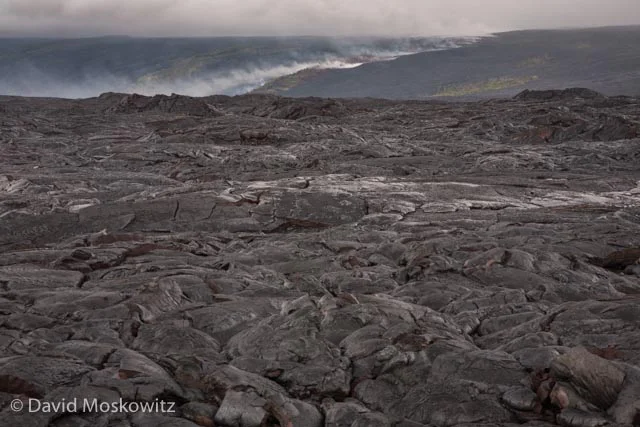The line of smoke in the distance marks the path of lava flowing down from the rift where it comes out of the earth, seen here across a vast plane of basalt from the recent flows from this rift which has been releasing lava on and off for years.
