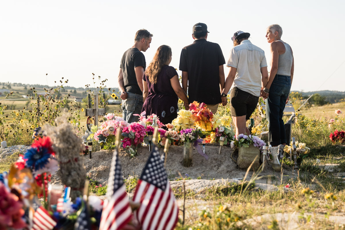 A moment of reflection and prayer at the graveyard at the Wounded Knee Massacre site. Adjacent to the mass grave from the massacre itself, others have subsequently been buried here. The victims of repeated waves of physical and cultural genecide including the massacre memorialized here, the complicated relationship between Lakota and other indigenous peoples and the United States is reflected in the American flags on the grave of tribal member on sovereign Lakota land.