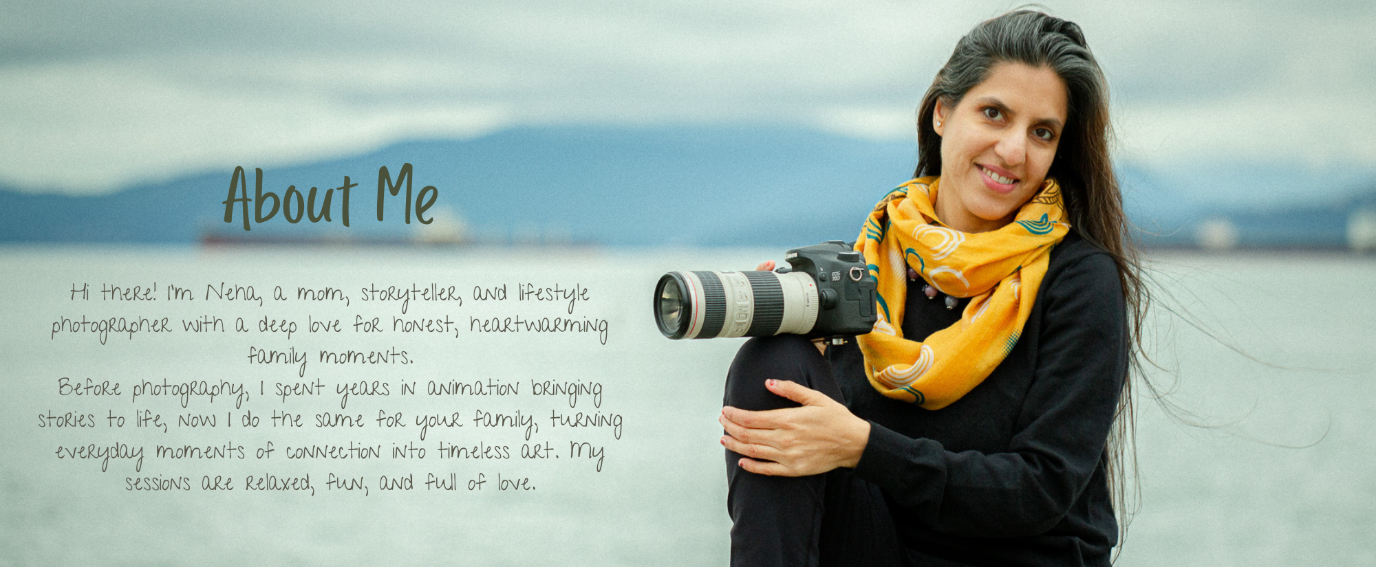 A woman with long dark hair wearing a yellow scarf and black jacket sitting outdoors near water, holding a camera with a large lens, smiling at the camera, with a mountain and cloudy sky in the background.