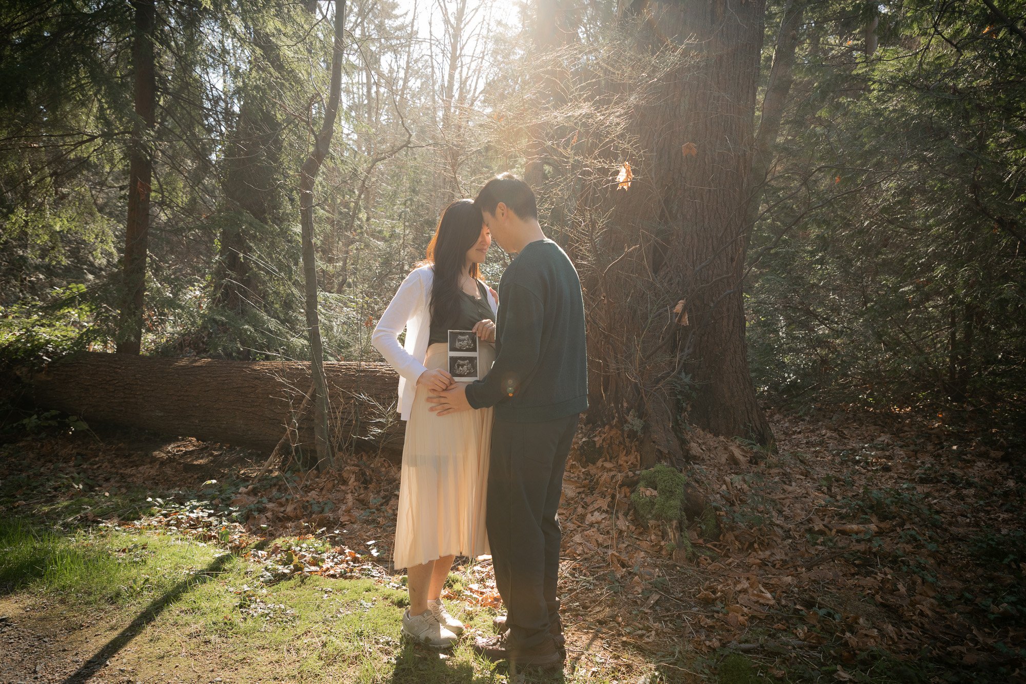 A couple standing close together in a sunlit forest, touching foreheads and holding ultrasound images, with fallen leaves on the ground and tall trees in the background.