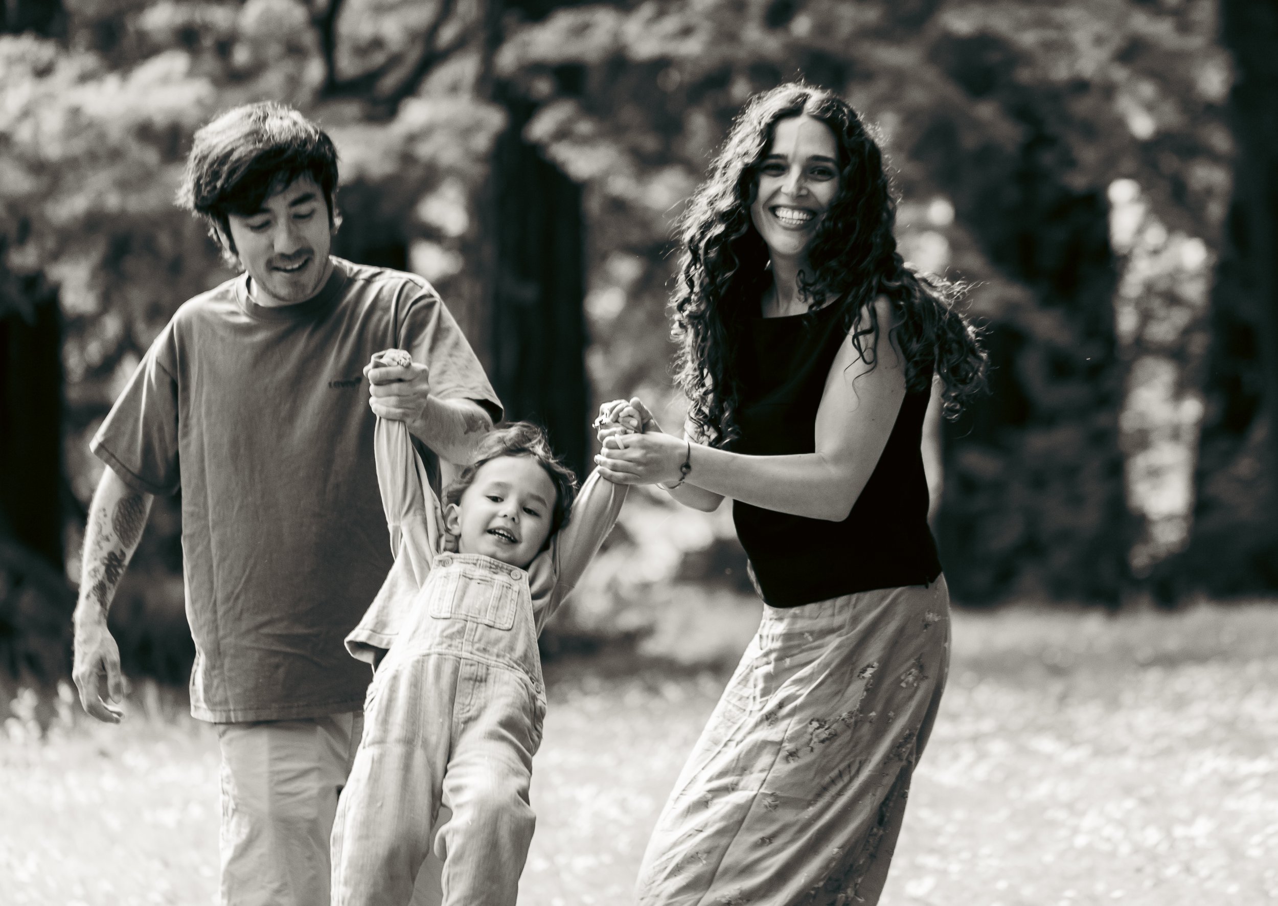 A family of three, a man, woman, and young girl, play together outdoors in a park with trees in the background. The woman and man are holding the girl by her hands and lifting her into the air, all smiling and enjoying the moment.
