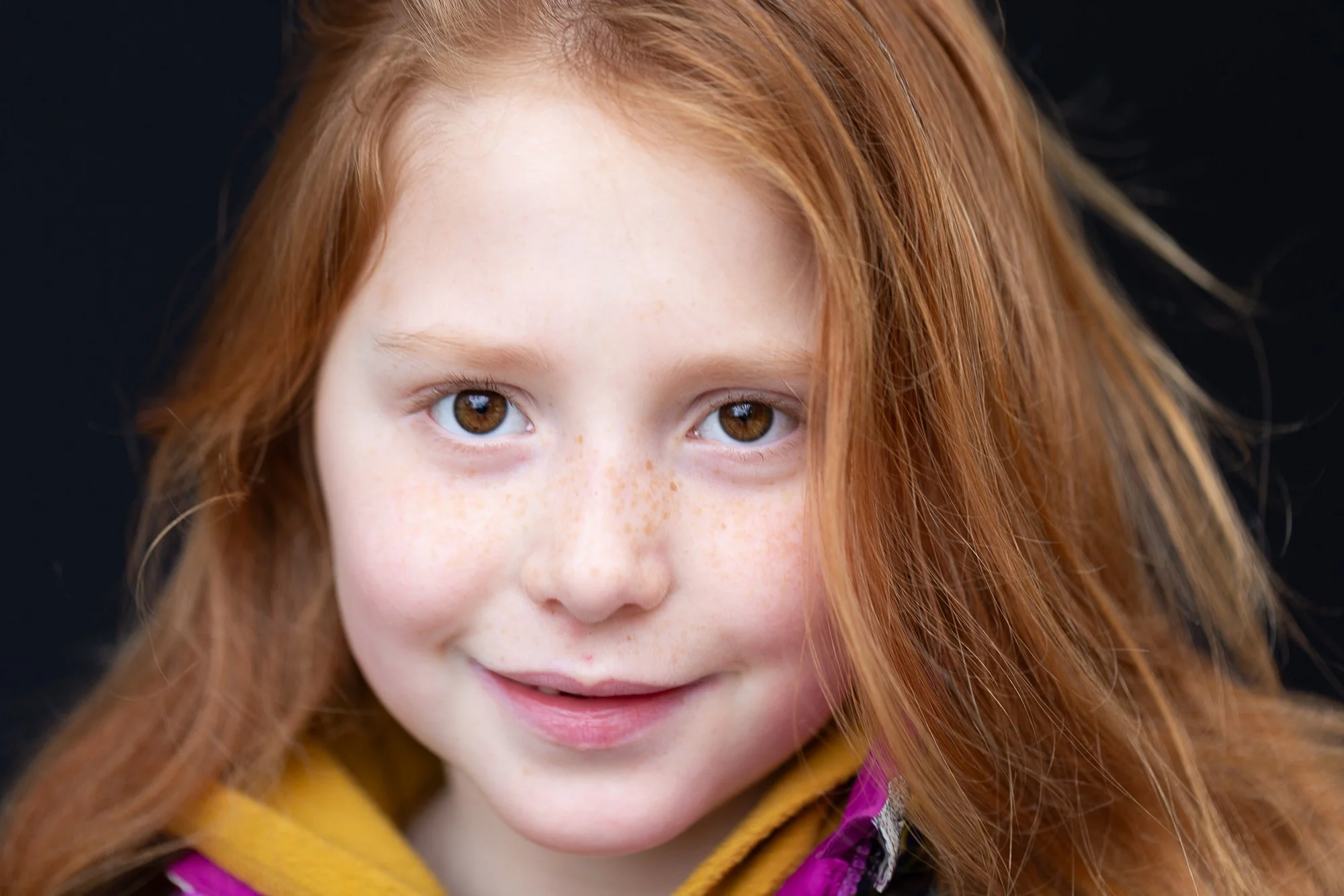 Close-up of a young girl with red hair, brown eyes, and freckles, smiling against a black background.