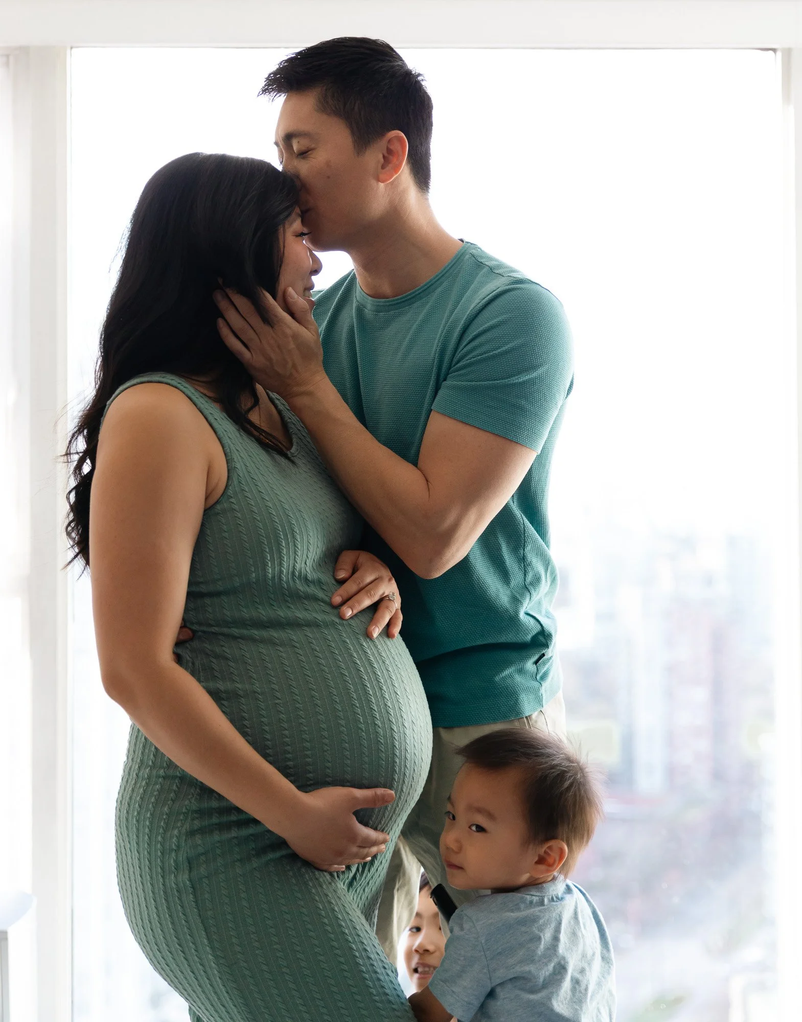 Pregnant woman with two young children and a man sharing a tender moment, with the man kissing the woman's forehead.