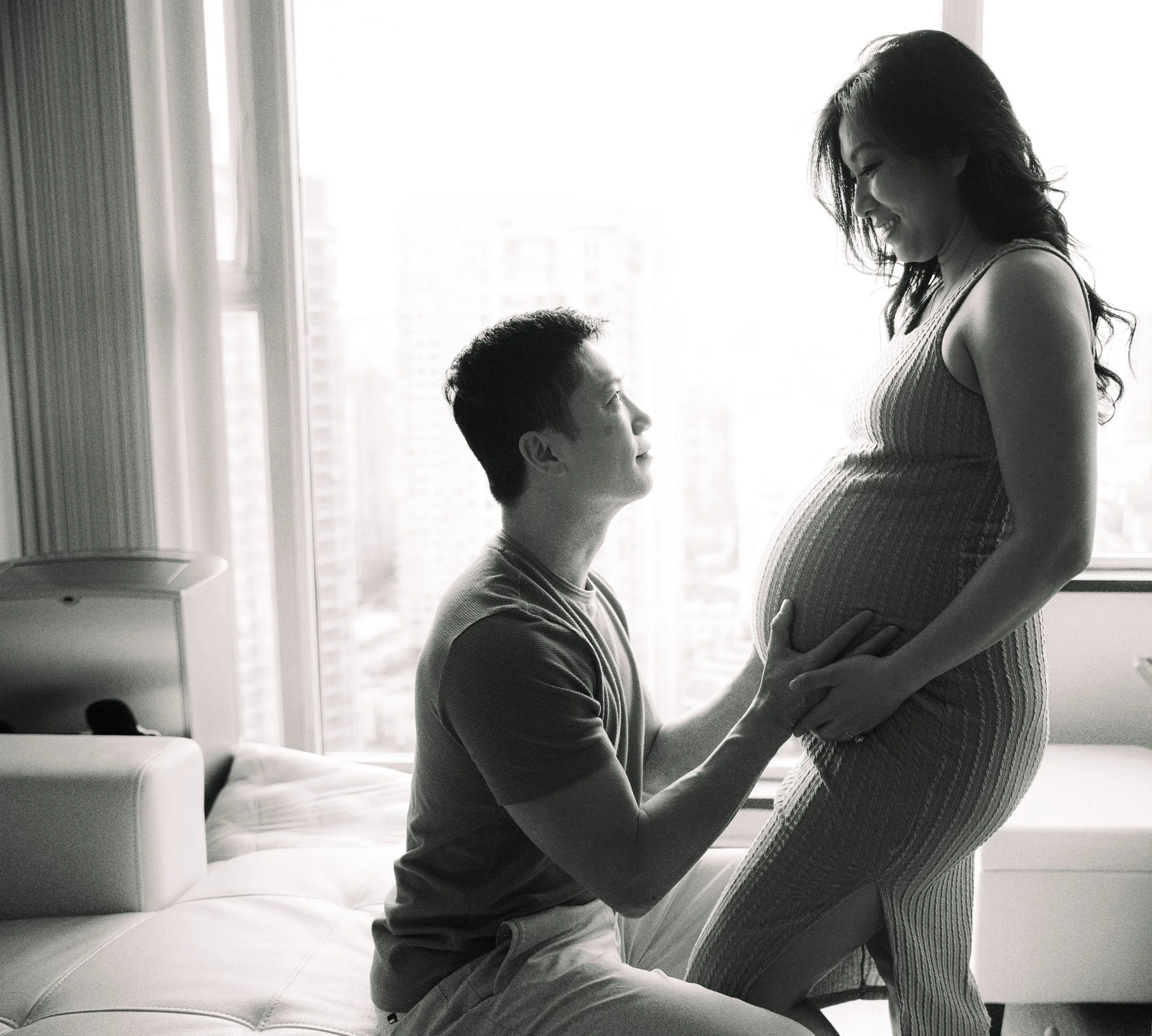 A man kneeling in front of a pregnant woman, holding her belly and gazing up at her, inside a room with large windows and city buildings in the background.