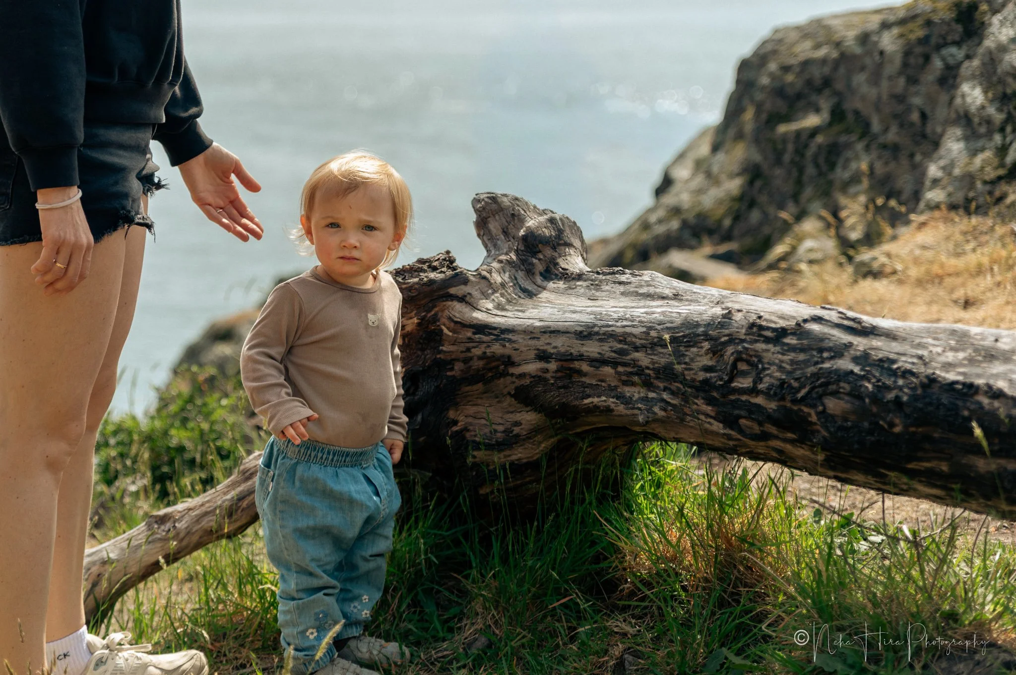 Toddler in tan top standing by driftwood log at Juniper Point, West Vancouver, with ocean in background