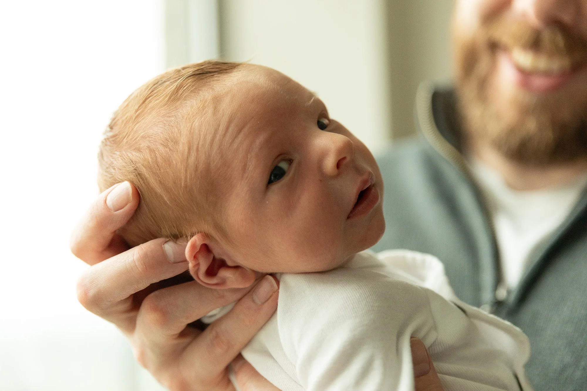 A close-up of a man holding a newborn baby with red hair and fair skin, the baby looking over his shoulder with a curious expression, both indoors near a window.