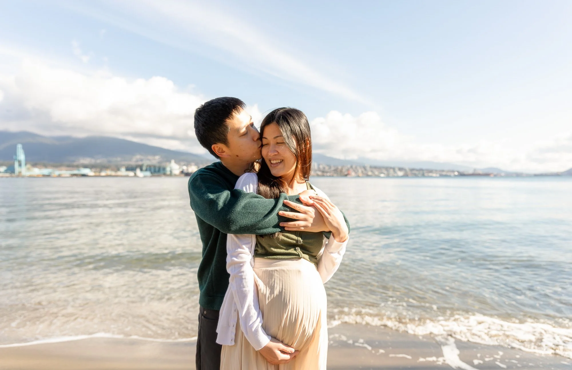 A couple standing on the beach, hugging and smiling with city skyline and mountains in the background.