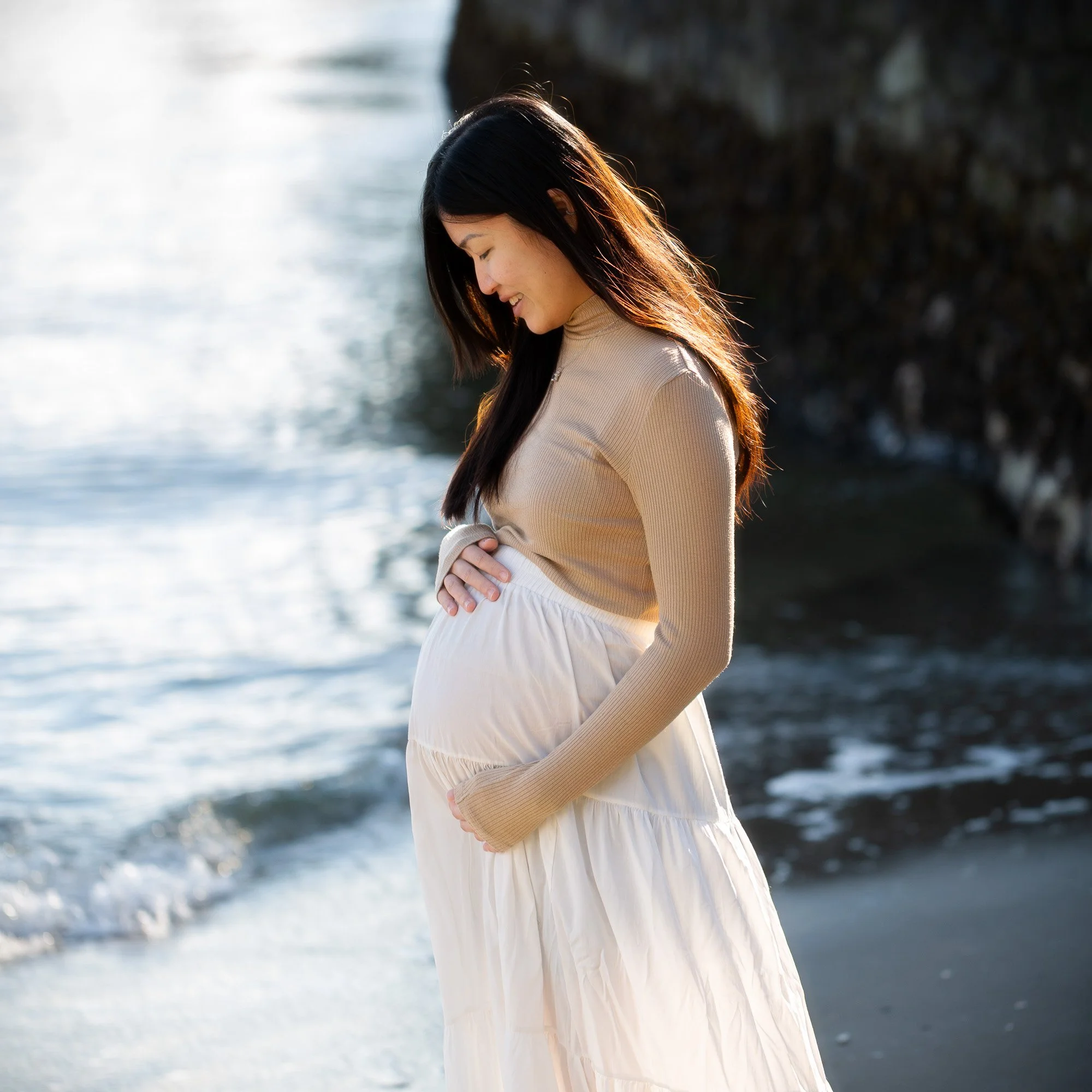 Pregnant woman standing on the beach with her hand on her belly, smiling while looking down, with ocean waves and rocks in the background.