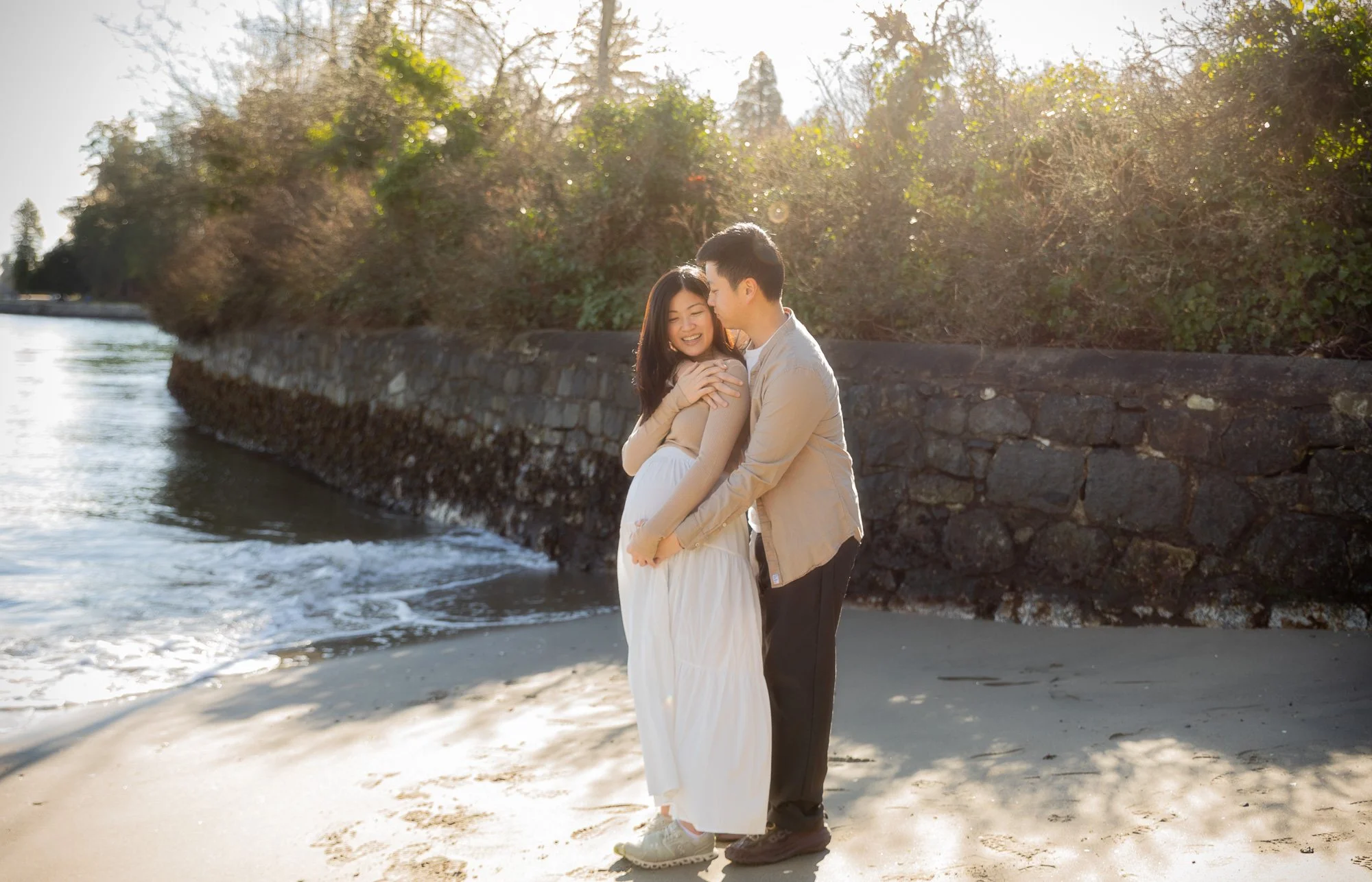 A couple embraces on a beach near a stone wall, with the woman smiling and the man gently kissing her forehead, during sunset.