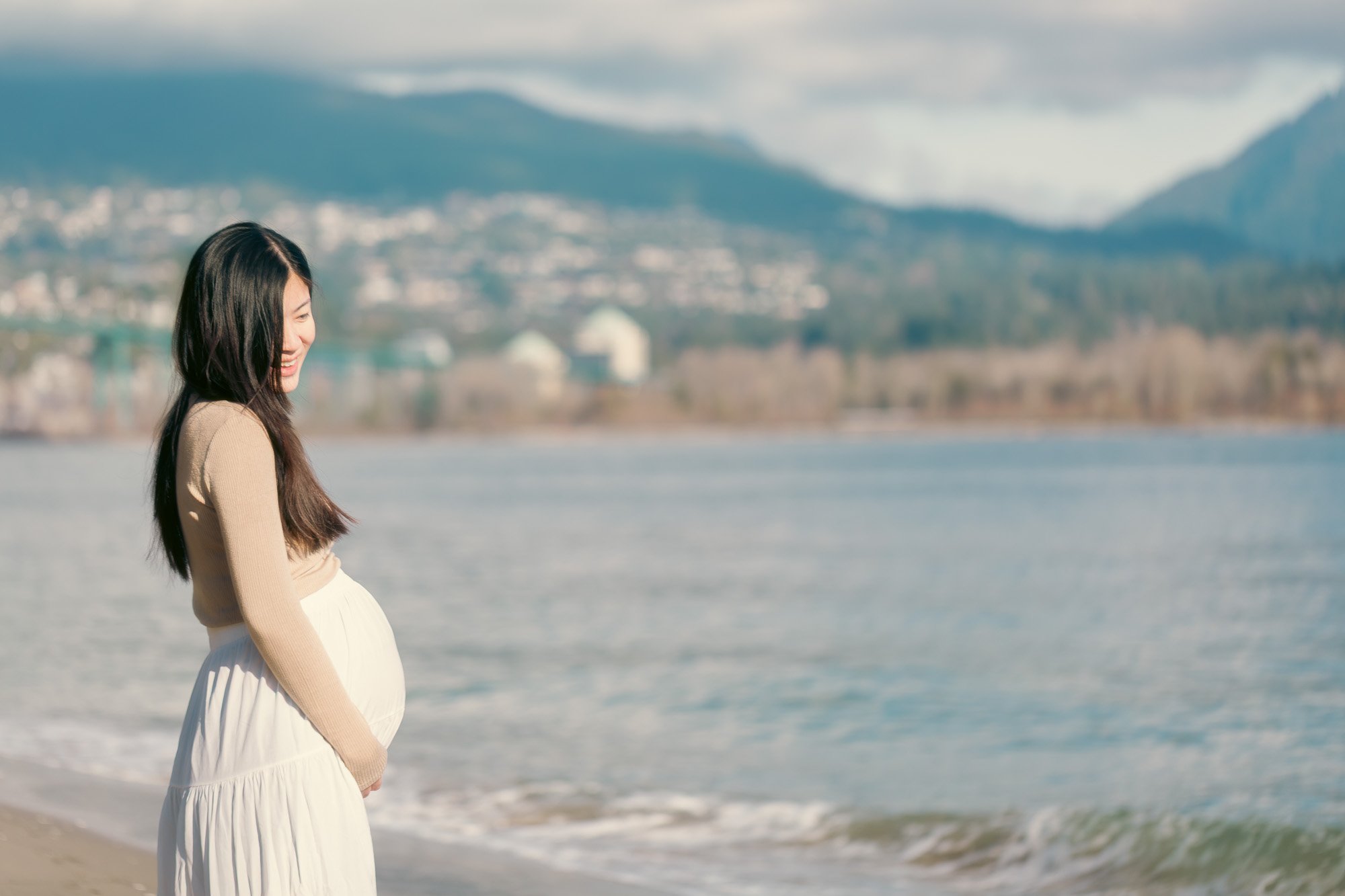 A pregnant woman in a beige sweater and white skirt stands on a beach near the water, smiling and looking down, with mountains and a town in the background.