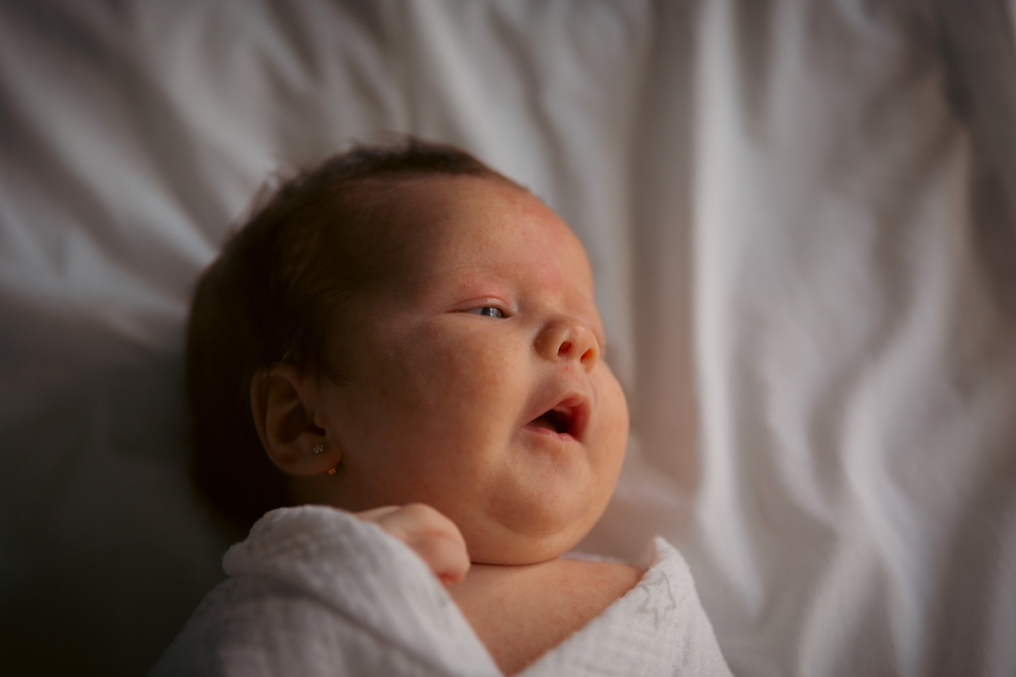 Close-up of a newborn baby lying on a white blanket, with skin redness and dark hair, looking to the side with slightly open mouth.