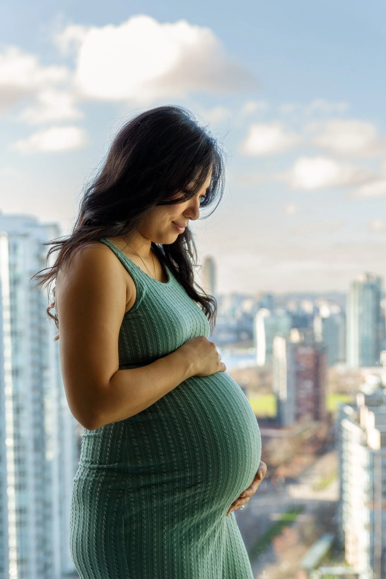 A pregnant woman in a green dress stands on a balcony with a cityscape in the background, holding her belly and looking down at it.