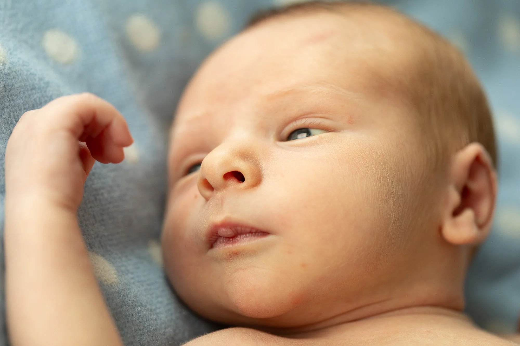 Close-up of a sleeping baby lying on a soft blanket with eyes half-open and hand near face.