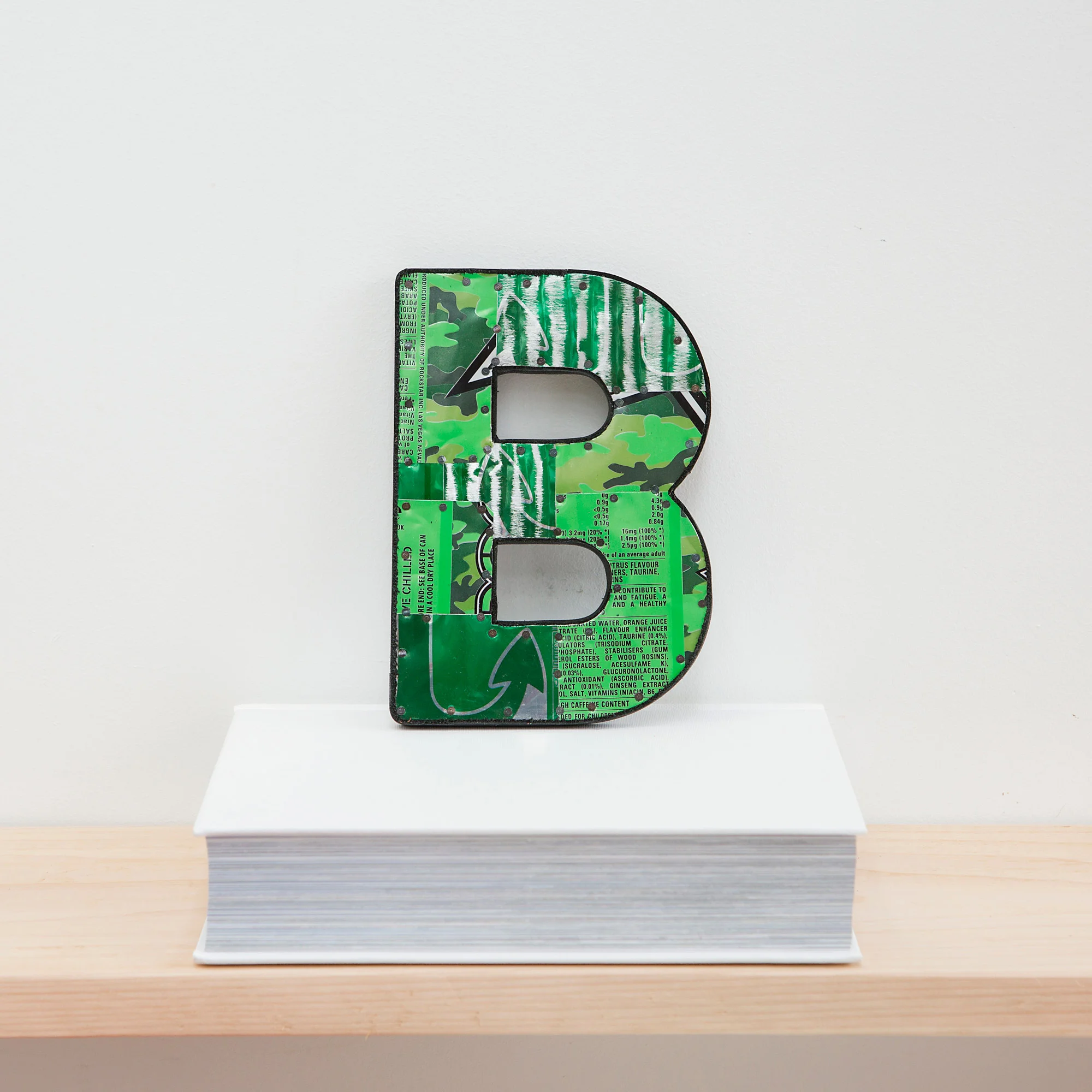 A green metal letter B font made from drinks can metal displayed on a white book , bespoke and handmade to order