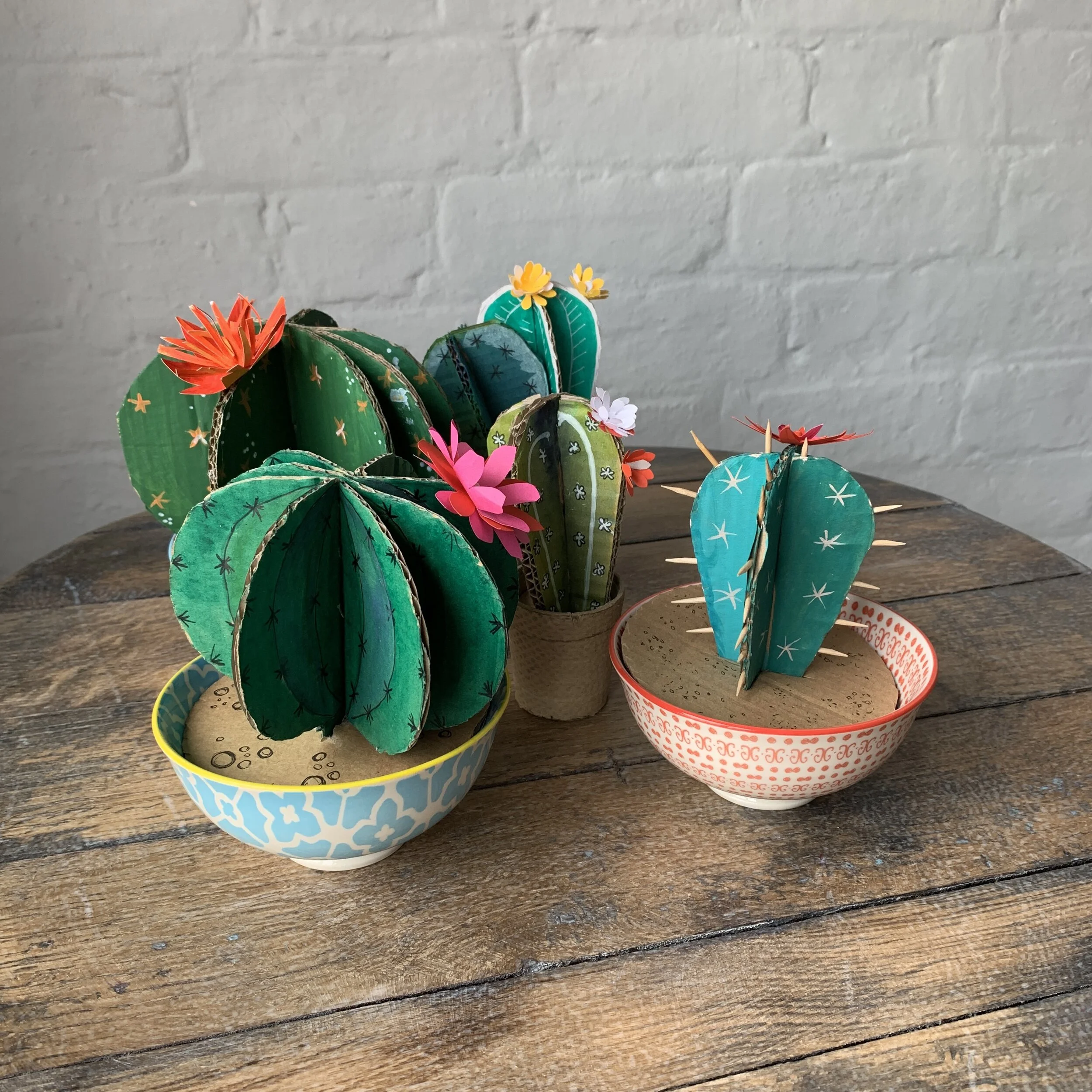 Several green craft cardboard barrel cacti on display n patterned bowls on a wooden table made from a card cactus making kit by Proteanart