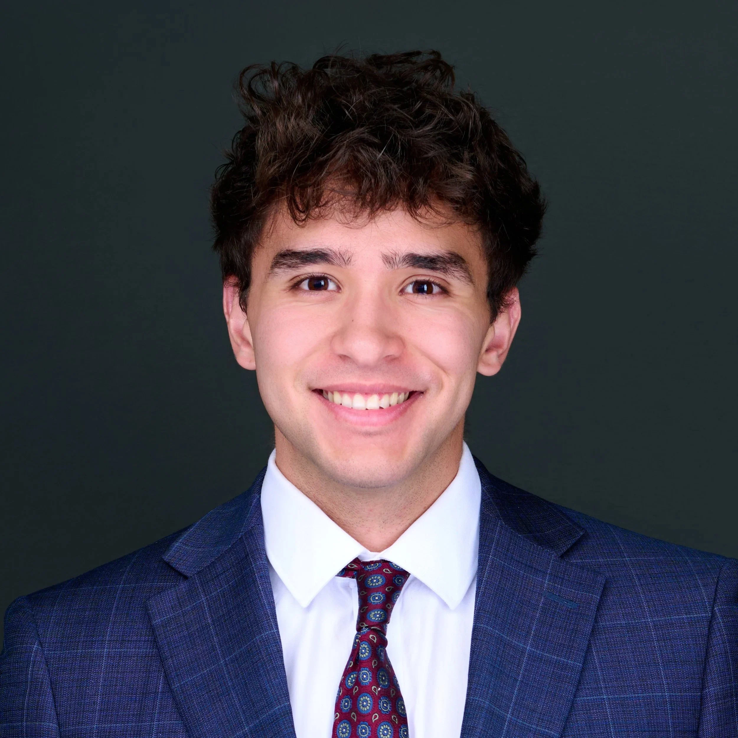 A young man with dark, curly hair wearing a navy blue suit, white shirt, and a patterned red tie, smiling against a dark backdrop.