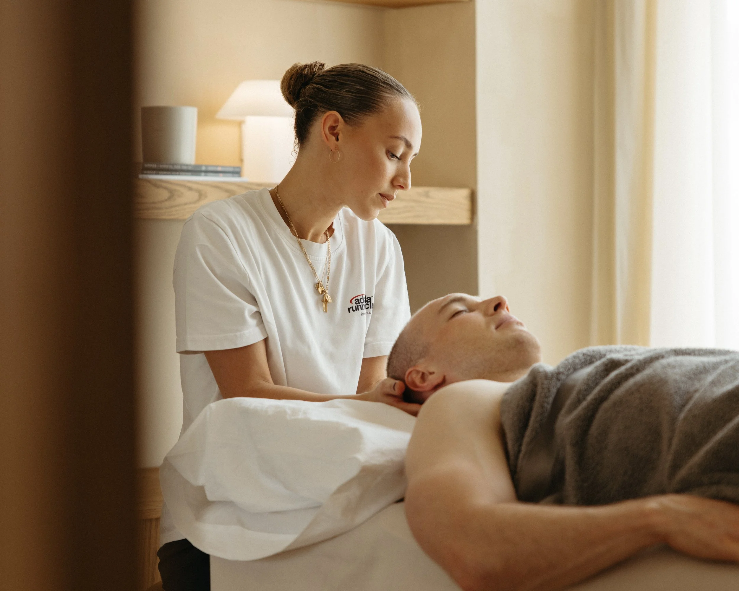 A woman with hair in a bun providing massage therapy to a man lying face up on a massage table, in a well-lit room.
