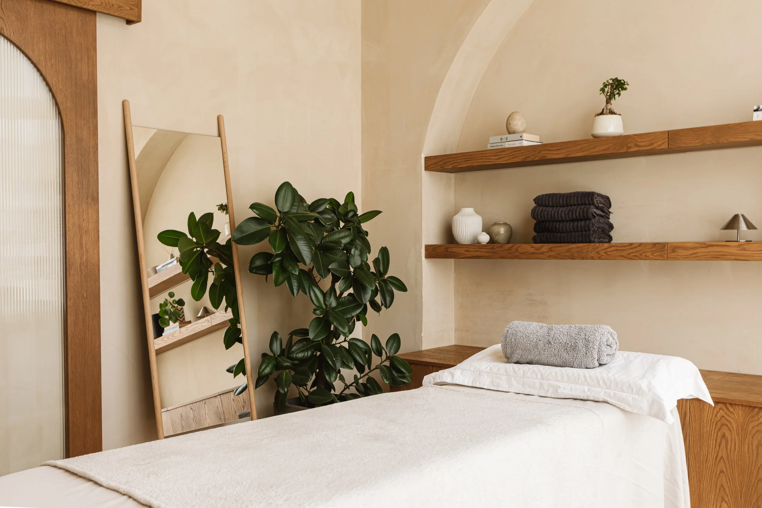 Interior of Nellie's clinic room with a massage table, rolled gray towel, plants, wooden shelves with decorative items, and a large mirror.