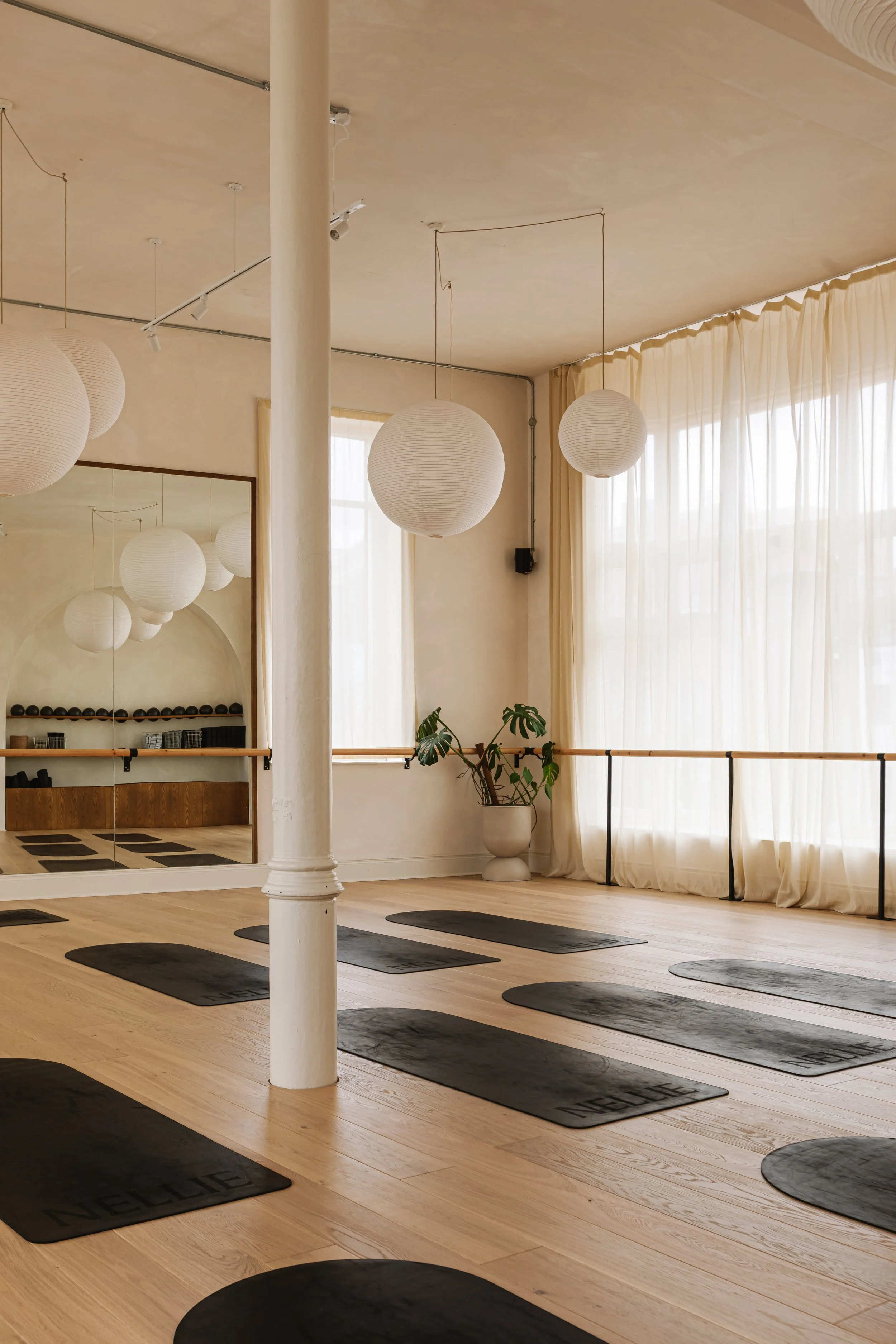 Yoga studio with black mats on wooden floor, large window with sheer curtains, hanging paper lanterns, and a mirror reflecting the room.