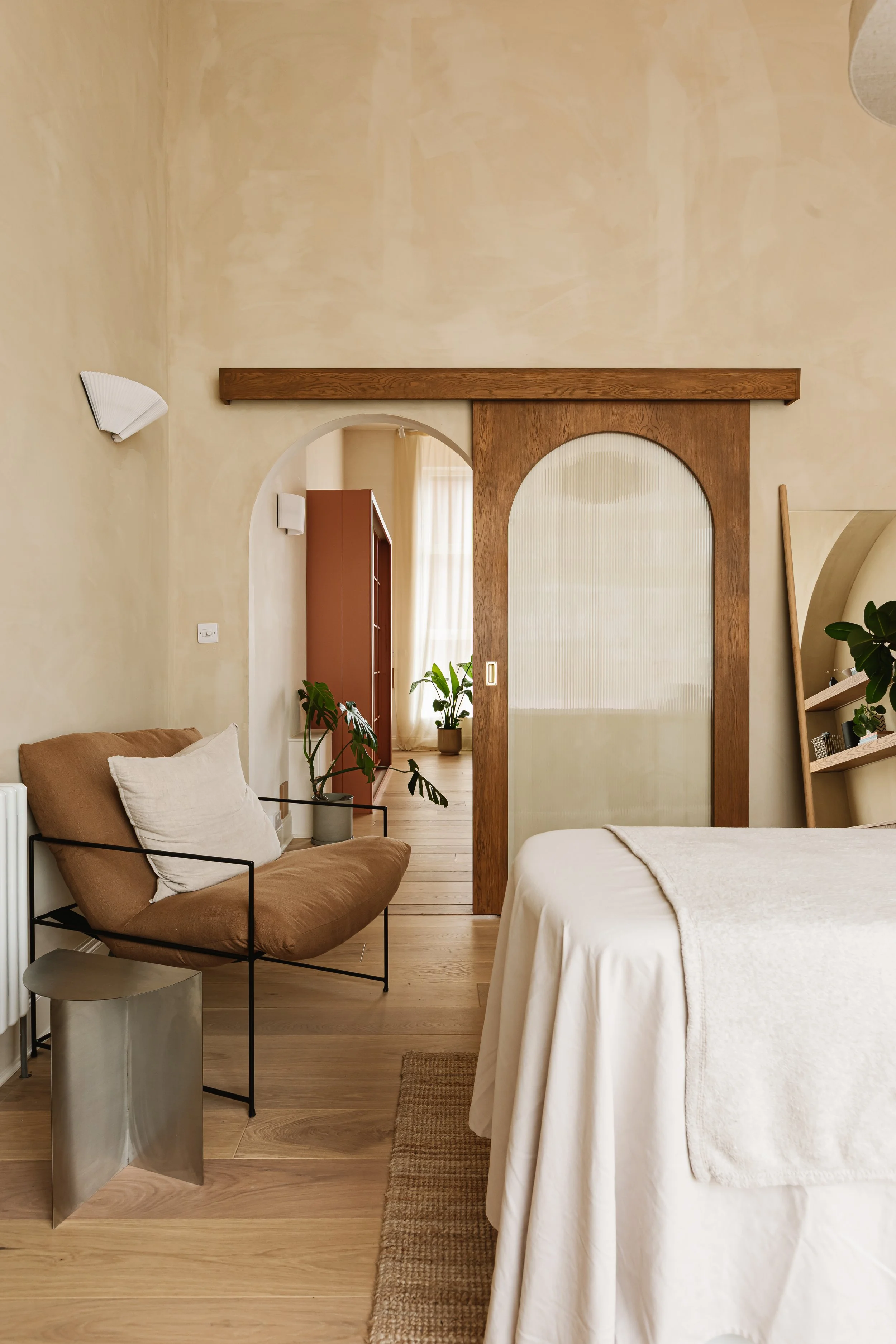 A bedroom with a beige wall, a bed with white bedding, a beige sofa with pillows, a metal side table, and a view through a wooden-framed archway into another room with plants and a curtain.