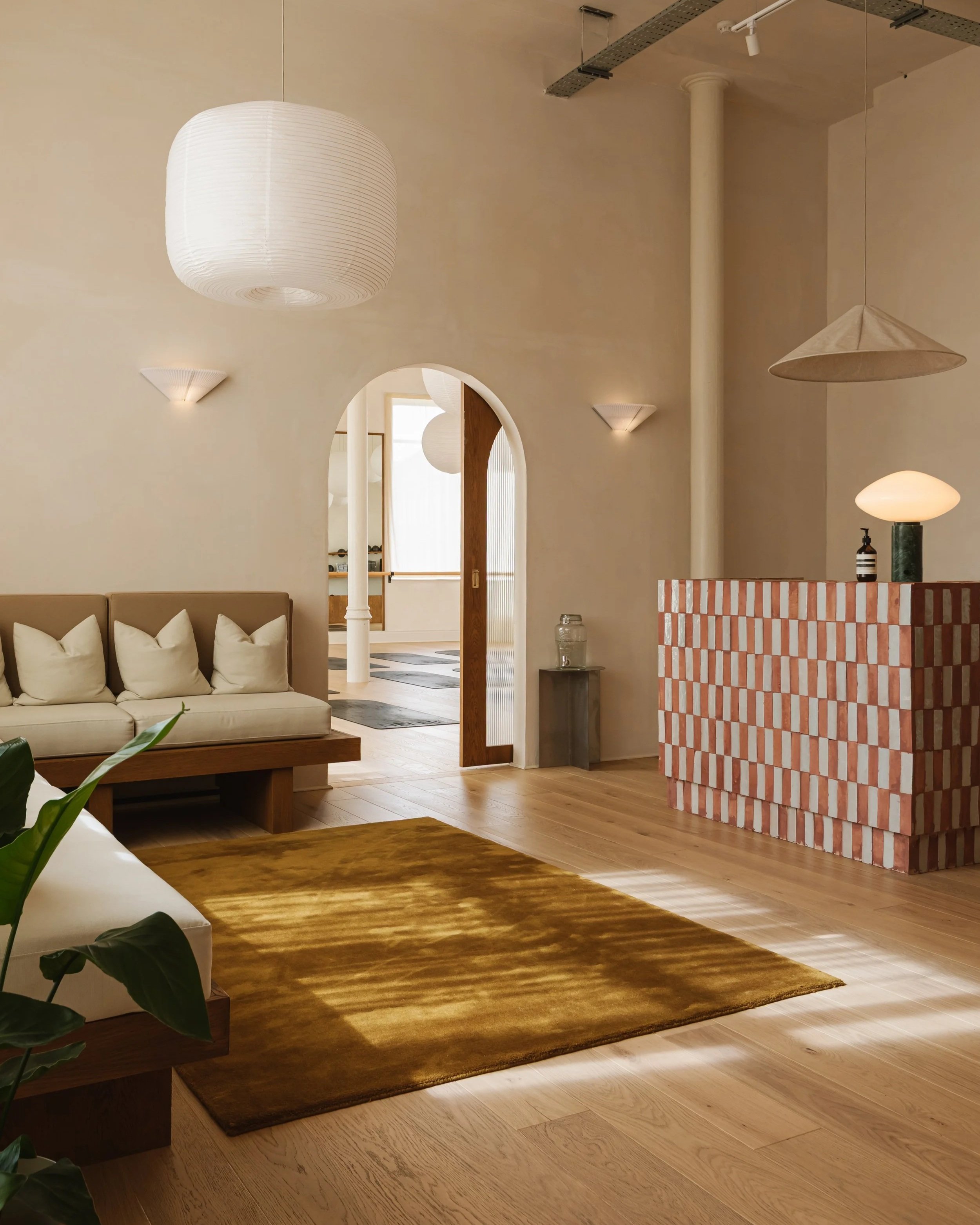 Interior of a modern room with light-colored wooden floors, a beige sofa with white pillows, a dark brown wooden table, a large mustard-colored rug, a white paper lantern ceiling light, wall-mounted lights, and a decorative divider with red and white tiles. There are indoor plants, a water dispenser, and stylish lamps.