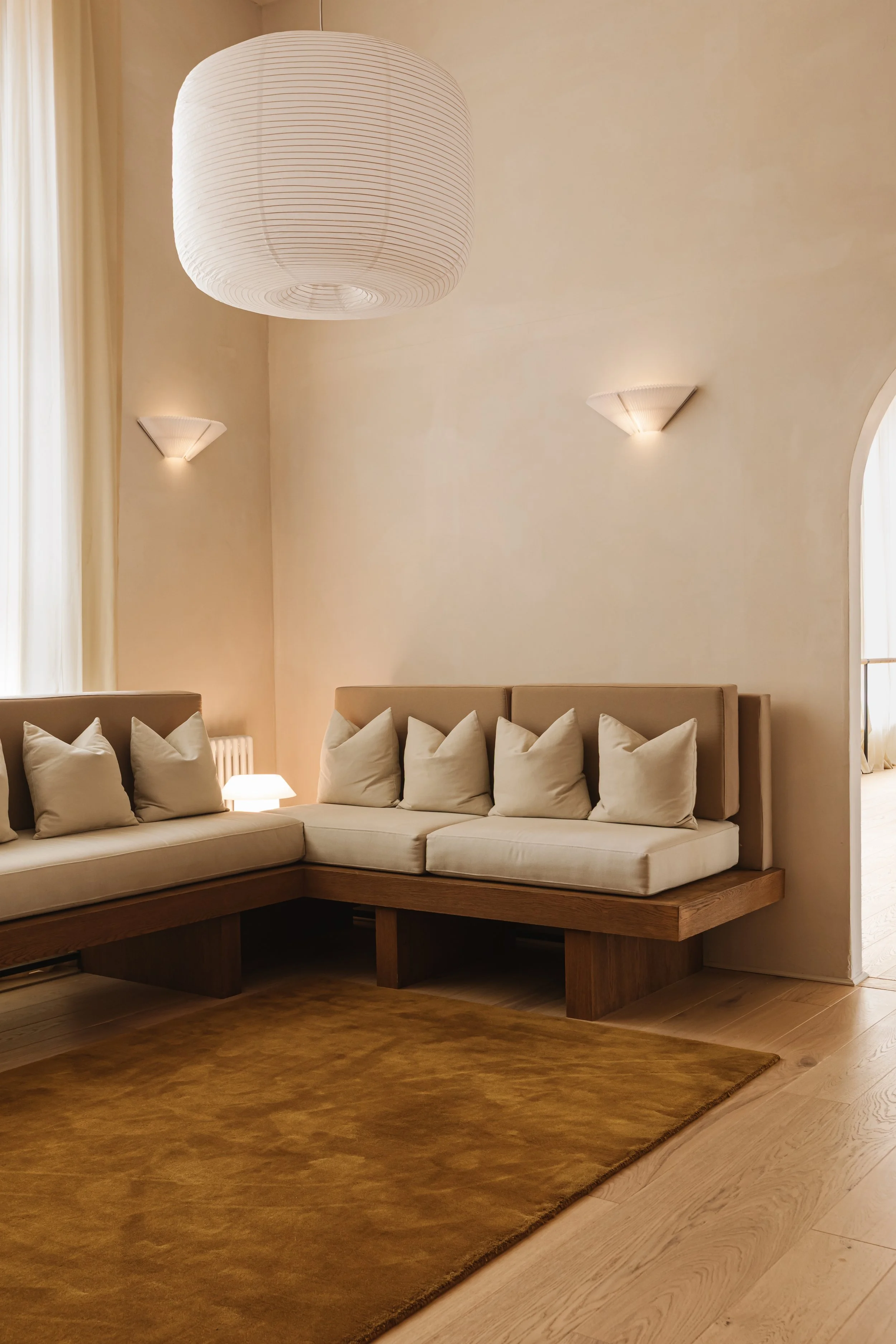 Living room corner with beige couch and white pillows, large paper lantern ceiling light, wall sconces, wooden floor, and brown area rug.