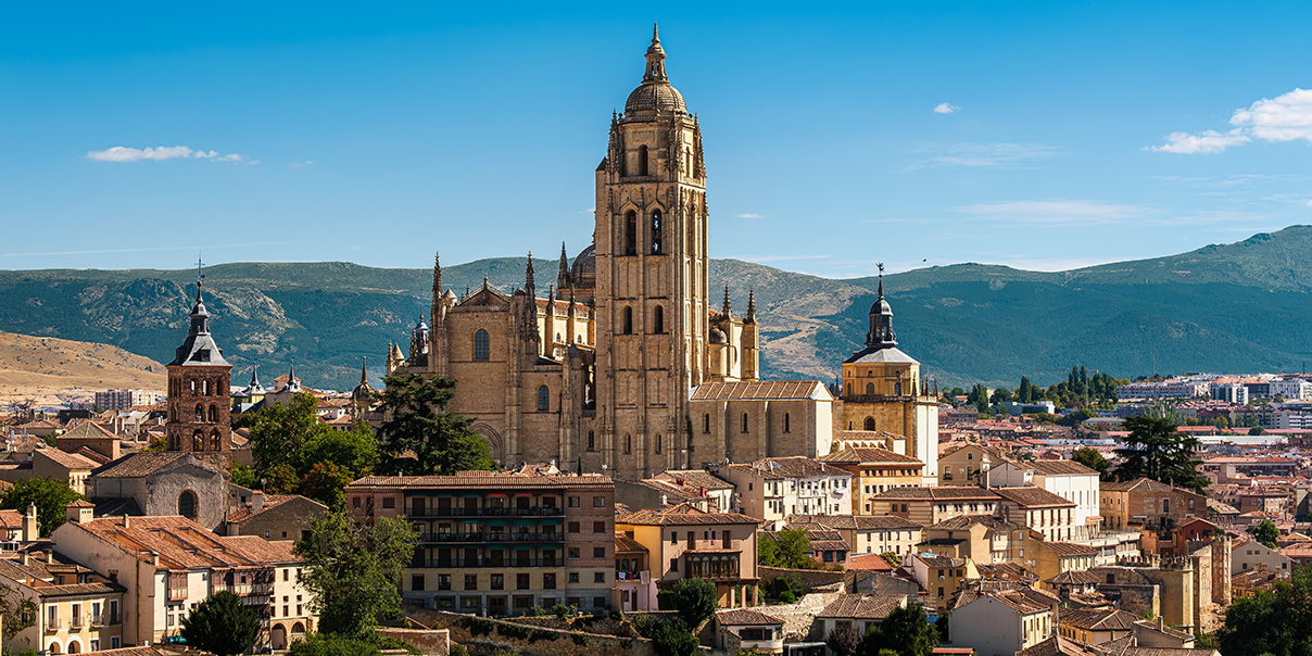 A stunning panoramic view of the Cathedral of Segovia rising majestically above the historic city skyline in Spain. Surrounded by traditional terracotta rooftops and backed by rolling mountains, this image highlights the grand scale and rich history 