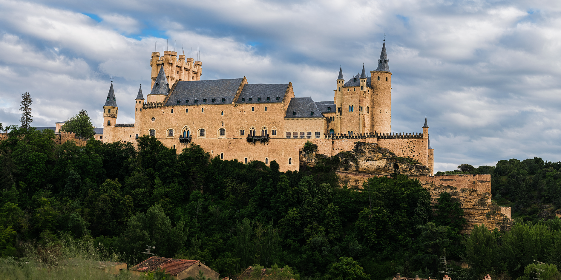 A breathtaking panoramic view of the Alcázar of Segovia rising high above the surrounding countryside in Spain. Captured from near the Iglesia de la Vera Cruz, this image showcases the impressive medieval turrets, golden stone walls, and dramatic ski