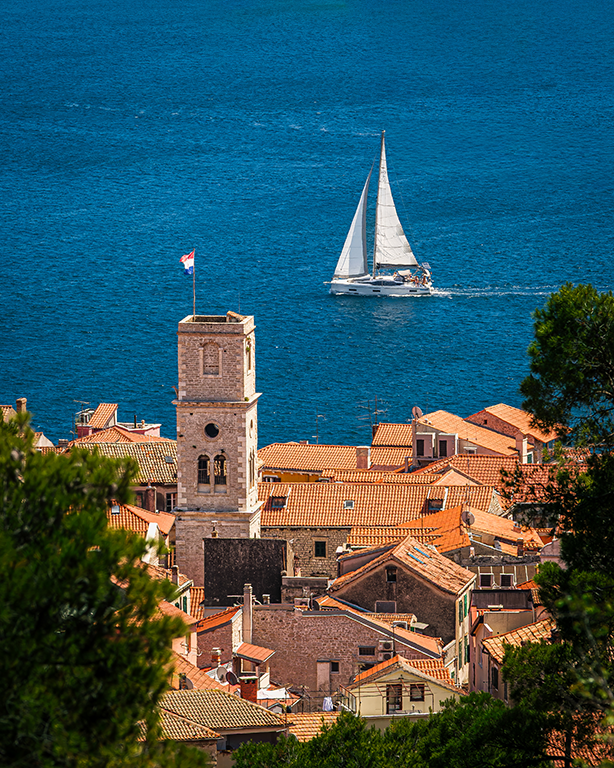 A high-angle view over the terracotta rooftops of Šibenik, Croatia, where a stone bell tower overlooks the deep blue Adriatic Sea. In the distance, a white sailboat glides past under clear Mediterranean skies. This print brings a classic, peaceful co