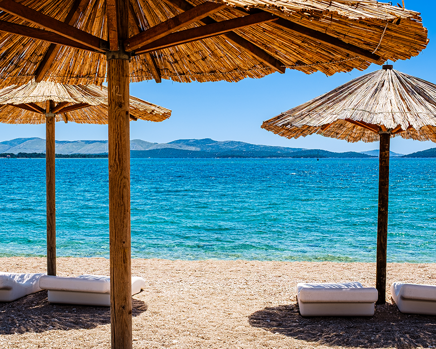 A vibrant, sun-soaked view of the Adriatic Sea along the Dalmatian Coast in Šibenik, Croatia. Rustic straw umbrellas frame the shimmering turquoise waters and distant coastal hills, capturing the quintessential Mediterranean beach day. Perfect for ad
