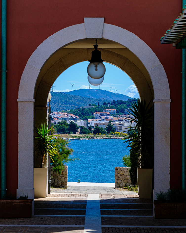 A beautifully framed perspective of the Adriatic Sea in Šibenik, Croatia, viewed through a terracotta and stone archway. A hanging globe lamp and lush potted greens complement the vivid contrast between shadow and bright Mediterranean light. Perfect 