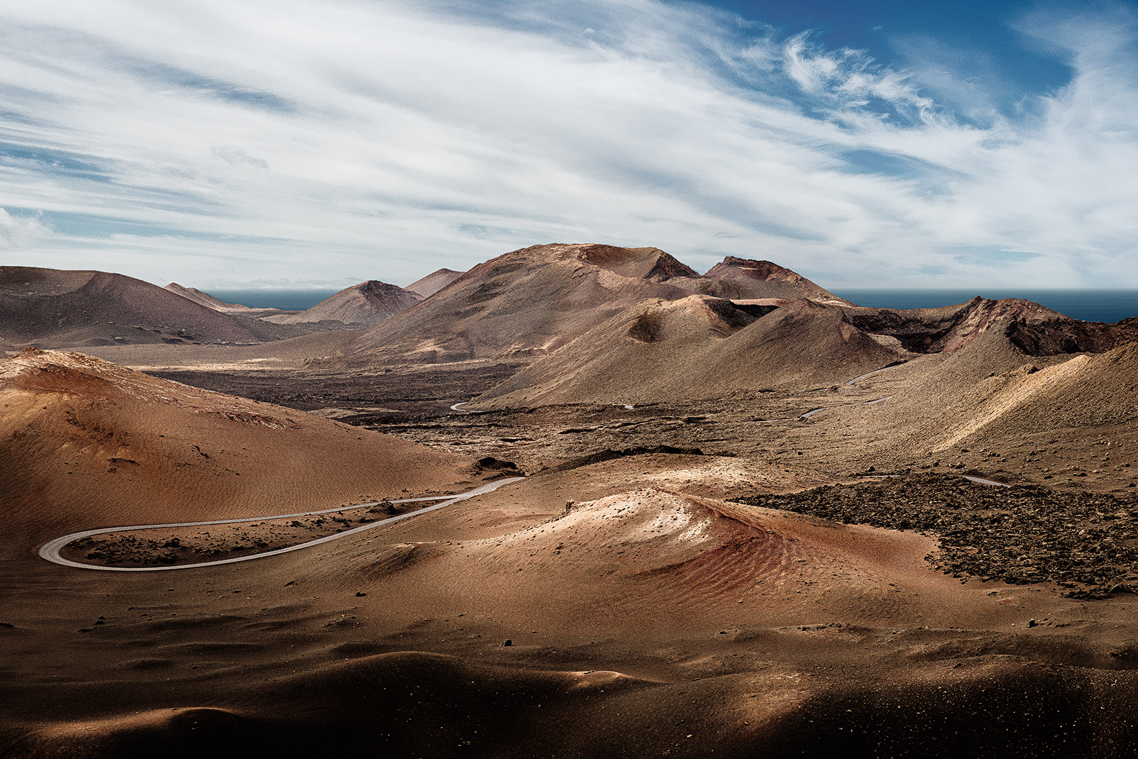 A panoramic view showcasing the stark, beautiful volcanic terrain of Timanfaya National Park in Lanzarote, Canary Islands. A solitary road snakes through layers of dark lava fields and ancient craters under a wide blue sky. This piece brings the dram