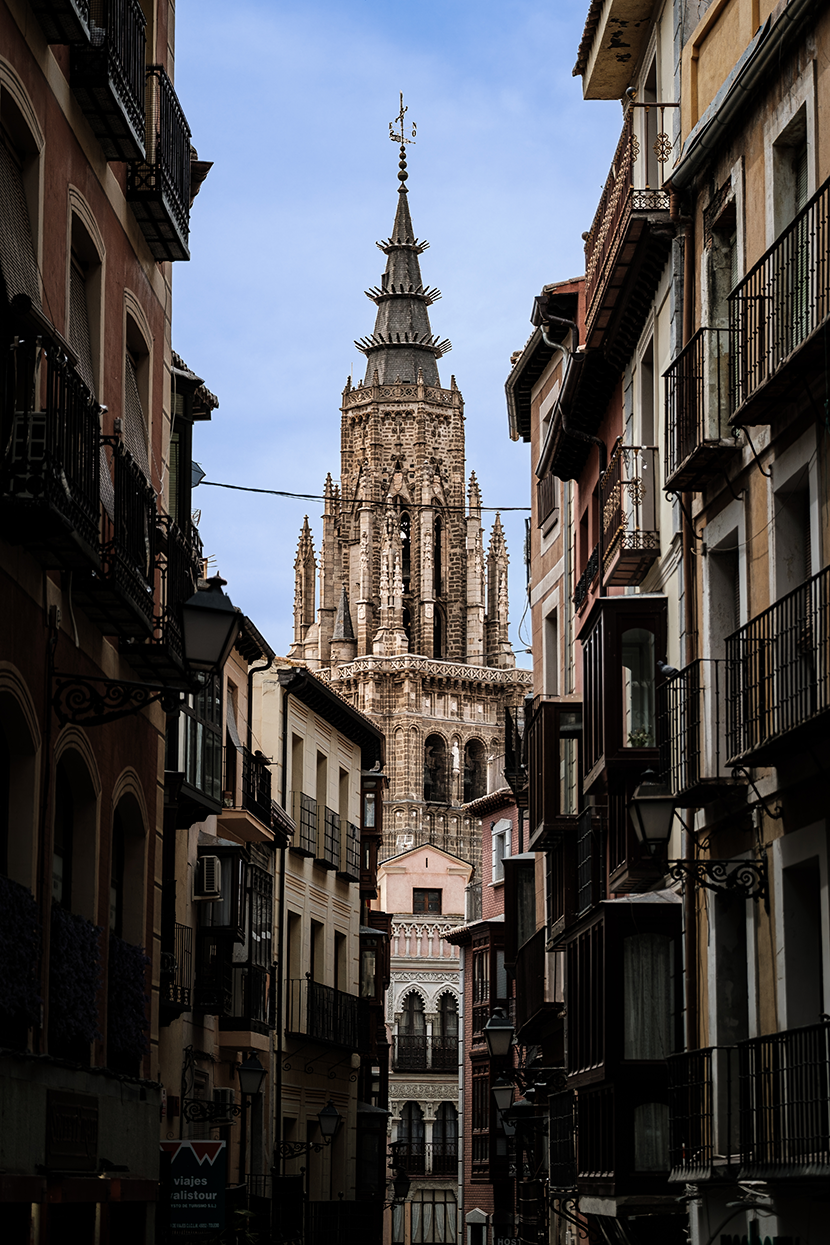 A dramatic vertical view of Toledo Cathedral’s intricate Gothic spire framed by the historic buildings and balconies of a narrow alleyway in Toledo, Spain. The clever composition creates a powerful sense of architectural depth and classic Old World a