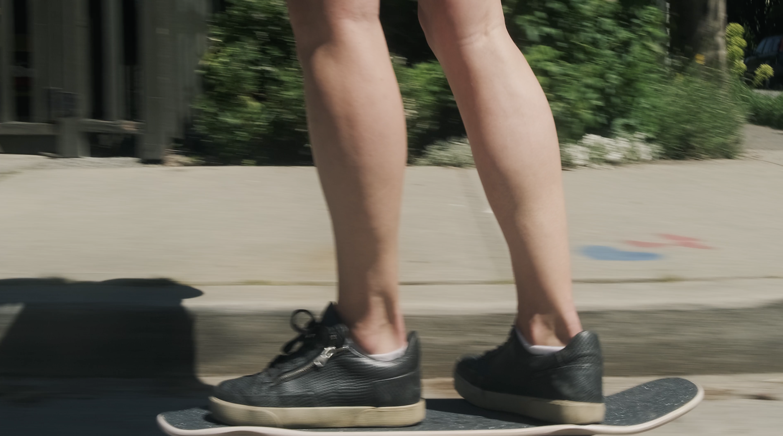 Close-up of a person's legs and feet riding a skateboard on a pavement, with green bushes and trees in the background.